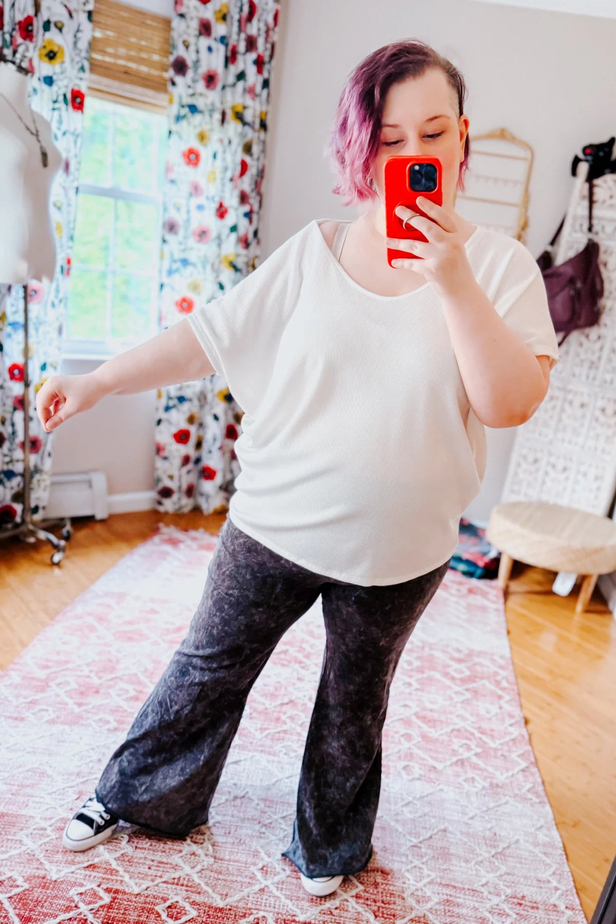 Woman taking mirror selfie wearing loose white top and ash black acid wash flare pants with sneakers indoors