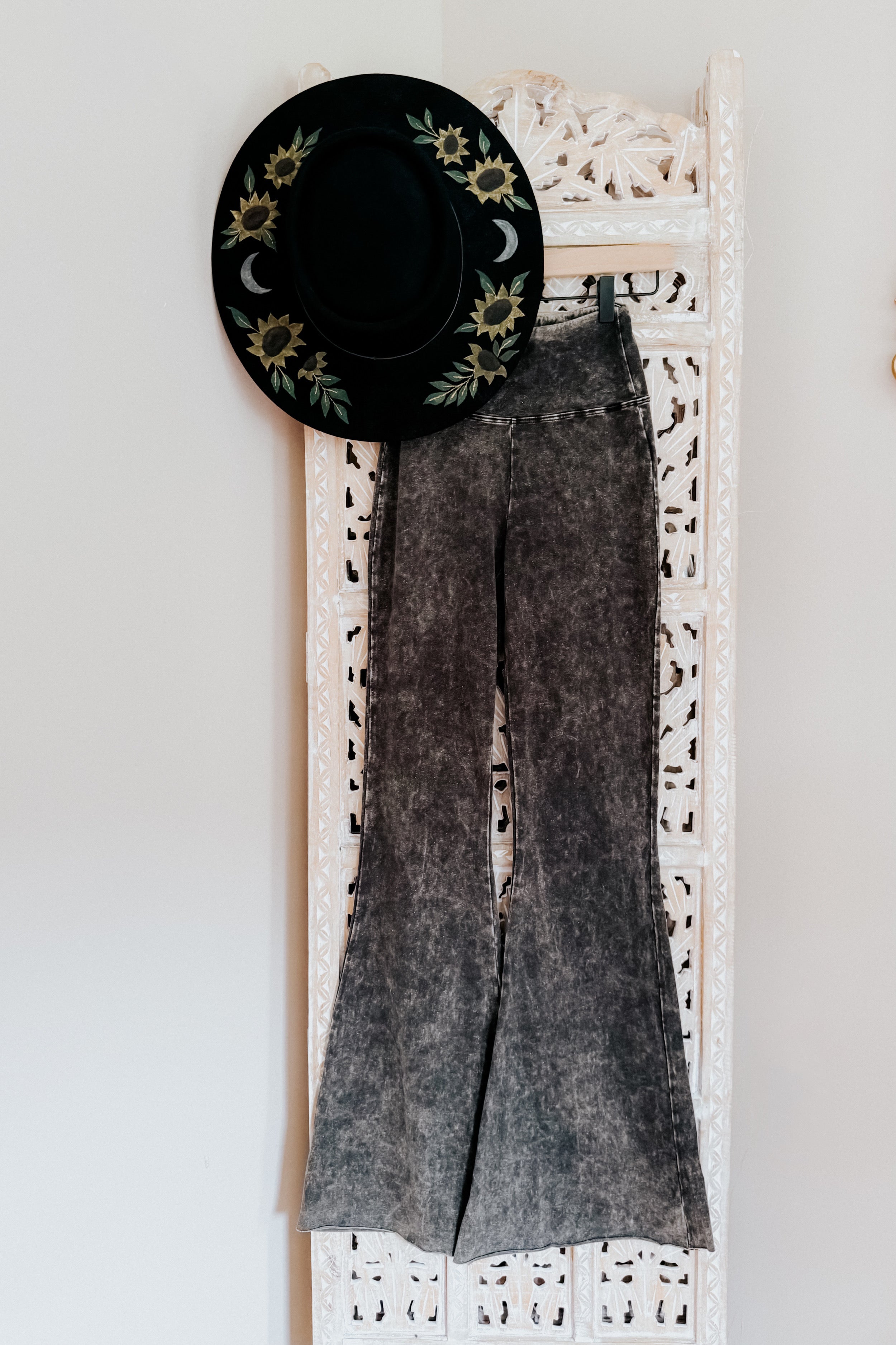 Ash black acid wash flare jeans hanging on carved white wooden room divider with black hat decorated with sunflowers and moons
