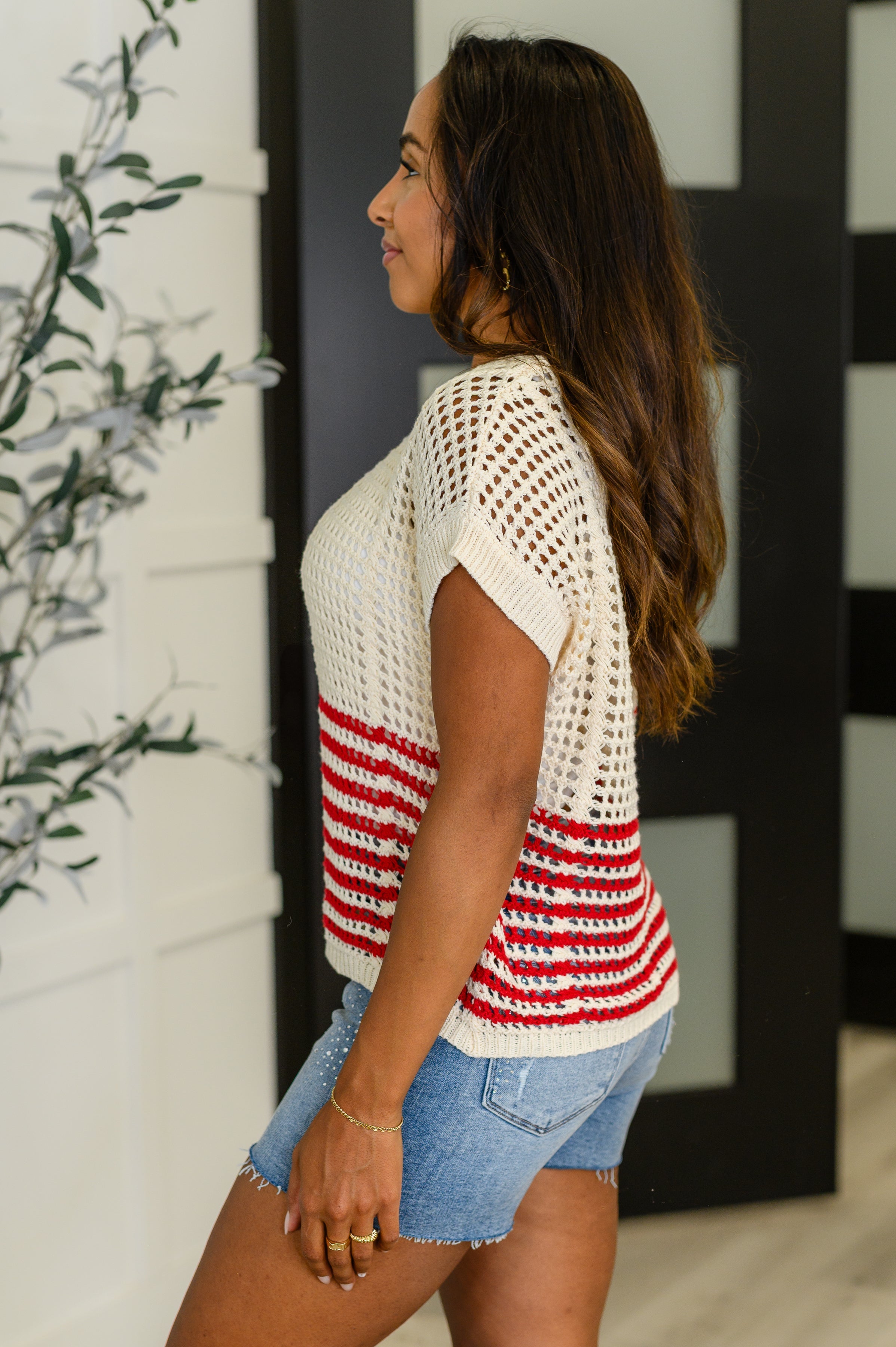 Woman wearing open-knit cream top with red stripes paired with frayed denim shorts in modern indoor setting