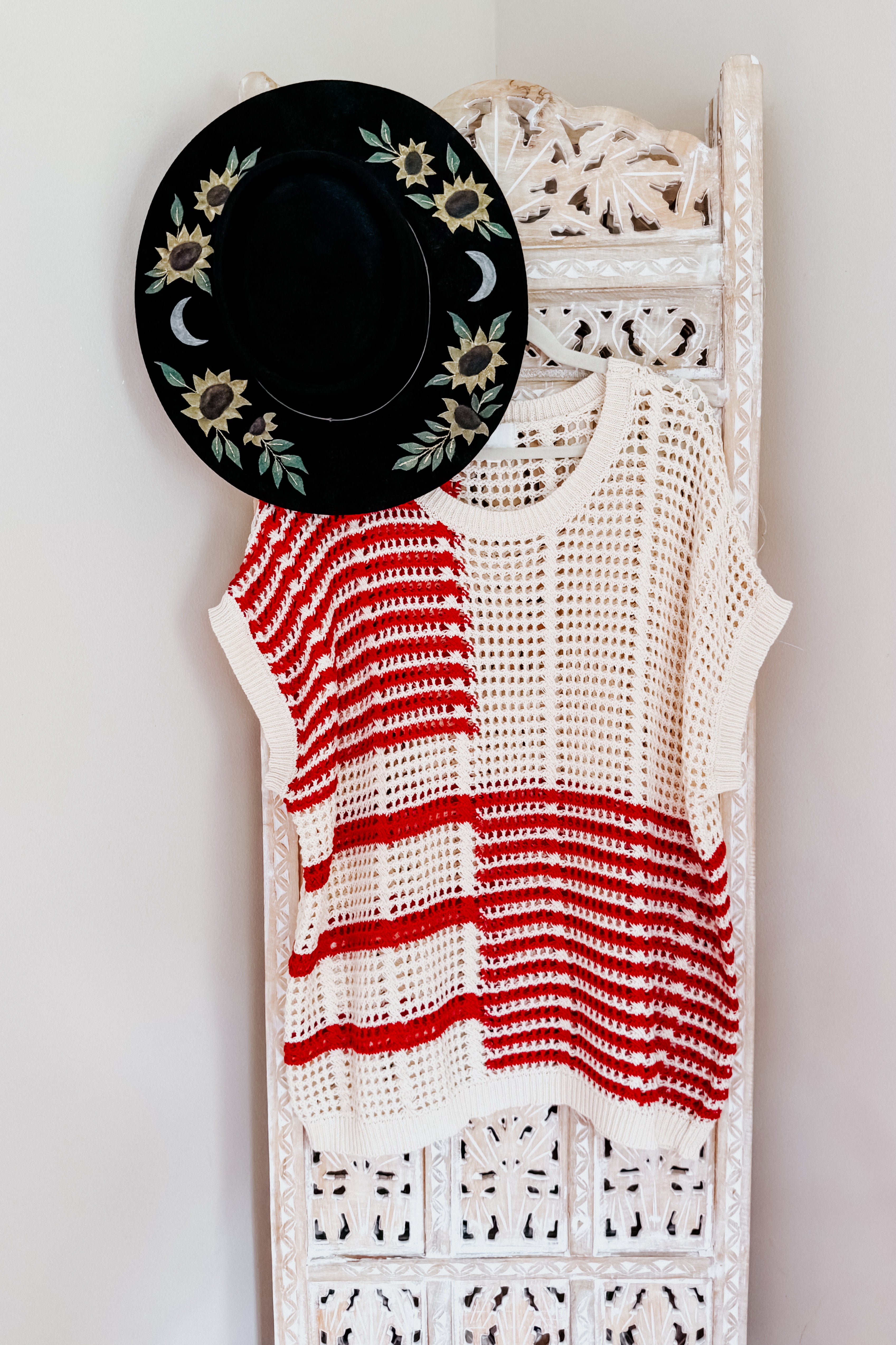 Crocheted open-knit top with bold red stripes hanging on carved white wooden screen with black floral hat above