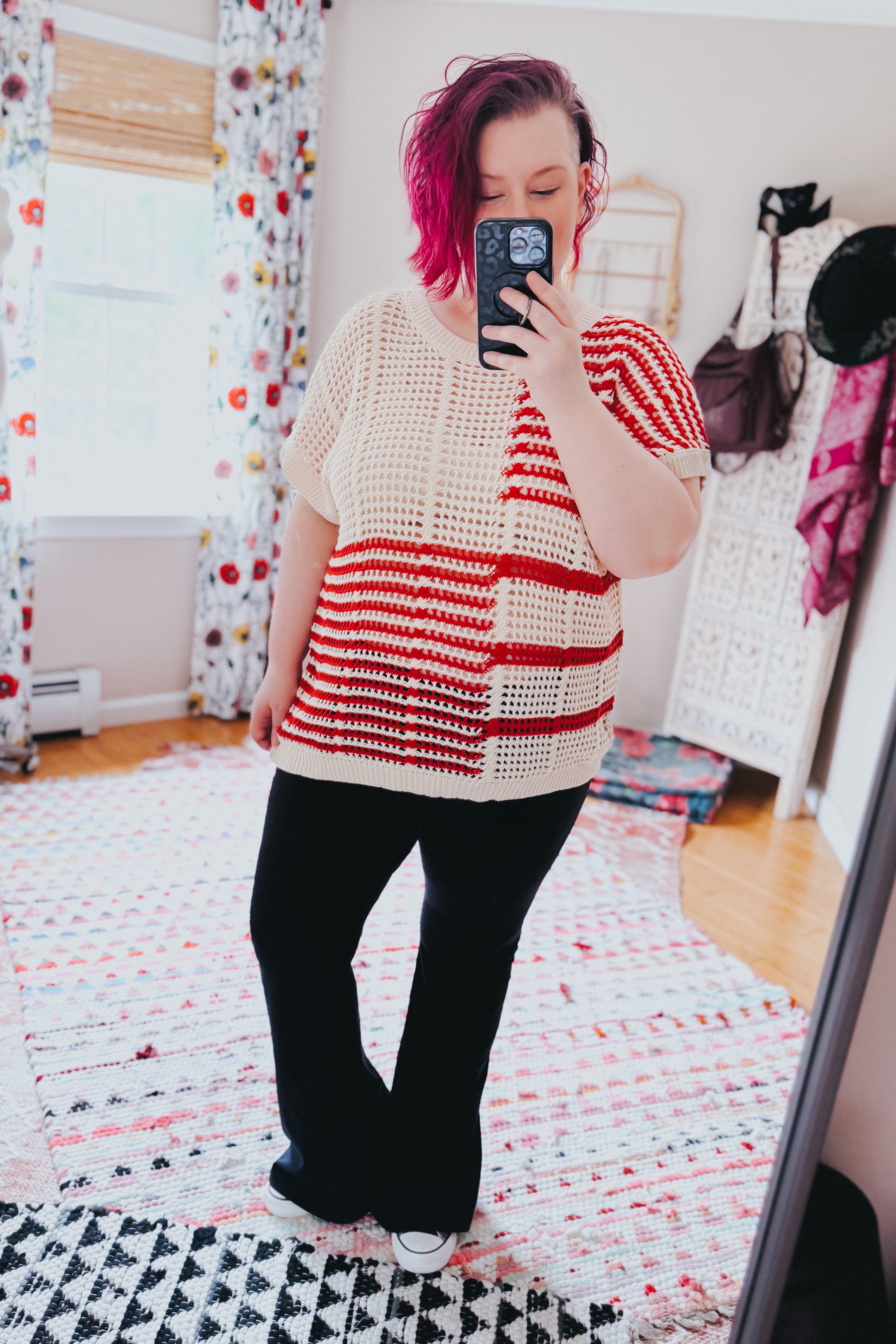 Woman with vibrant magenta hair wearing red striped net tee and black pants taking mirror selfie in cozy bright room
