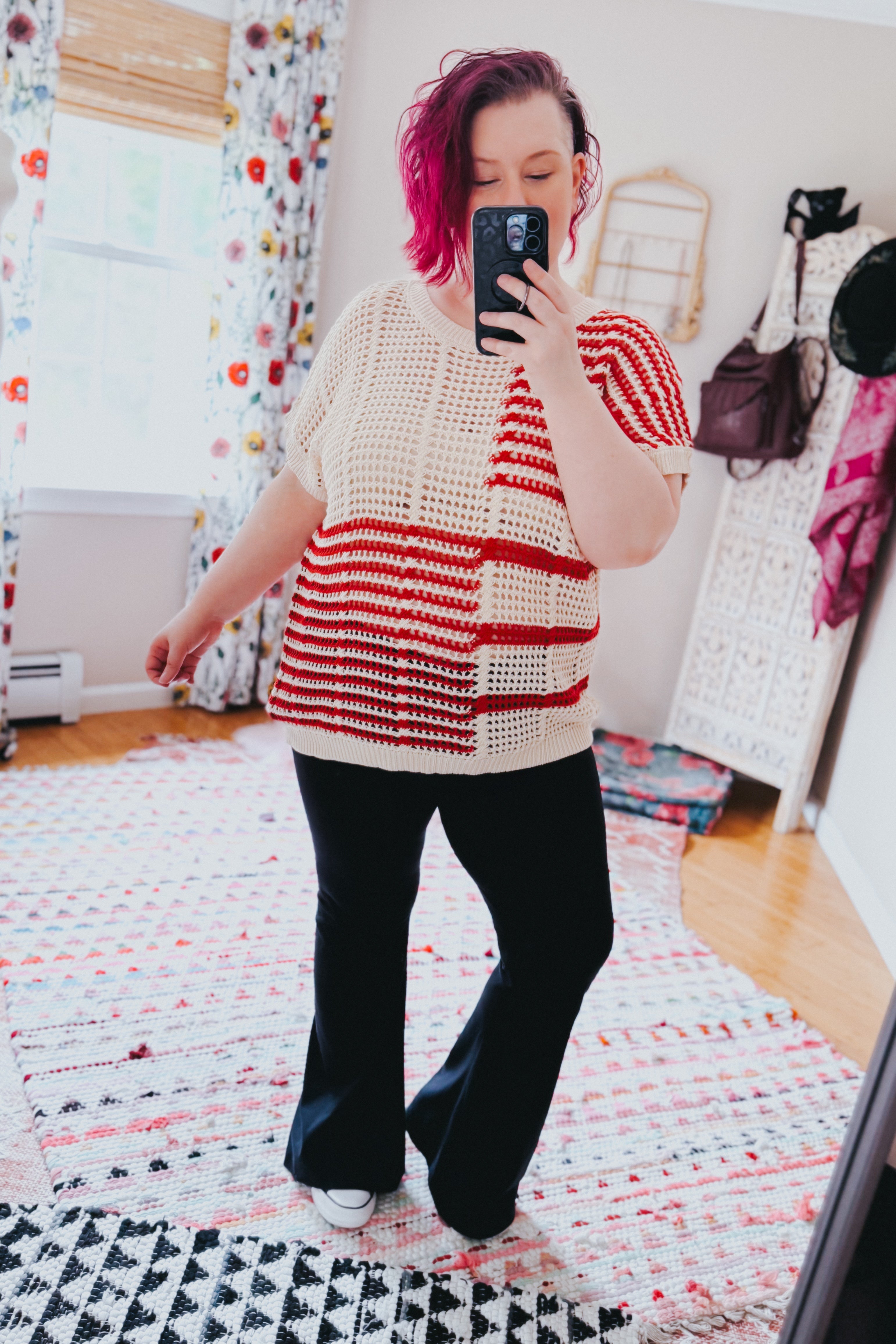 Woman with magenta hair wearing cream and red striped net tee with black flare pants taking mirror selfie in cozy room