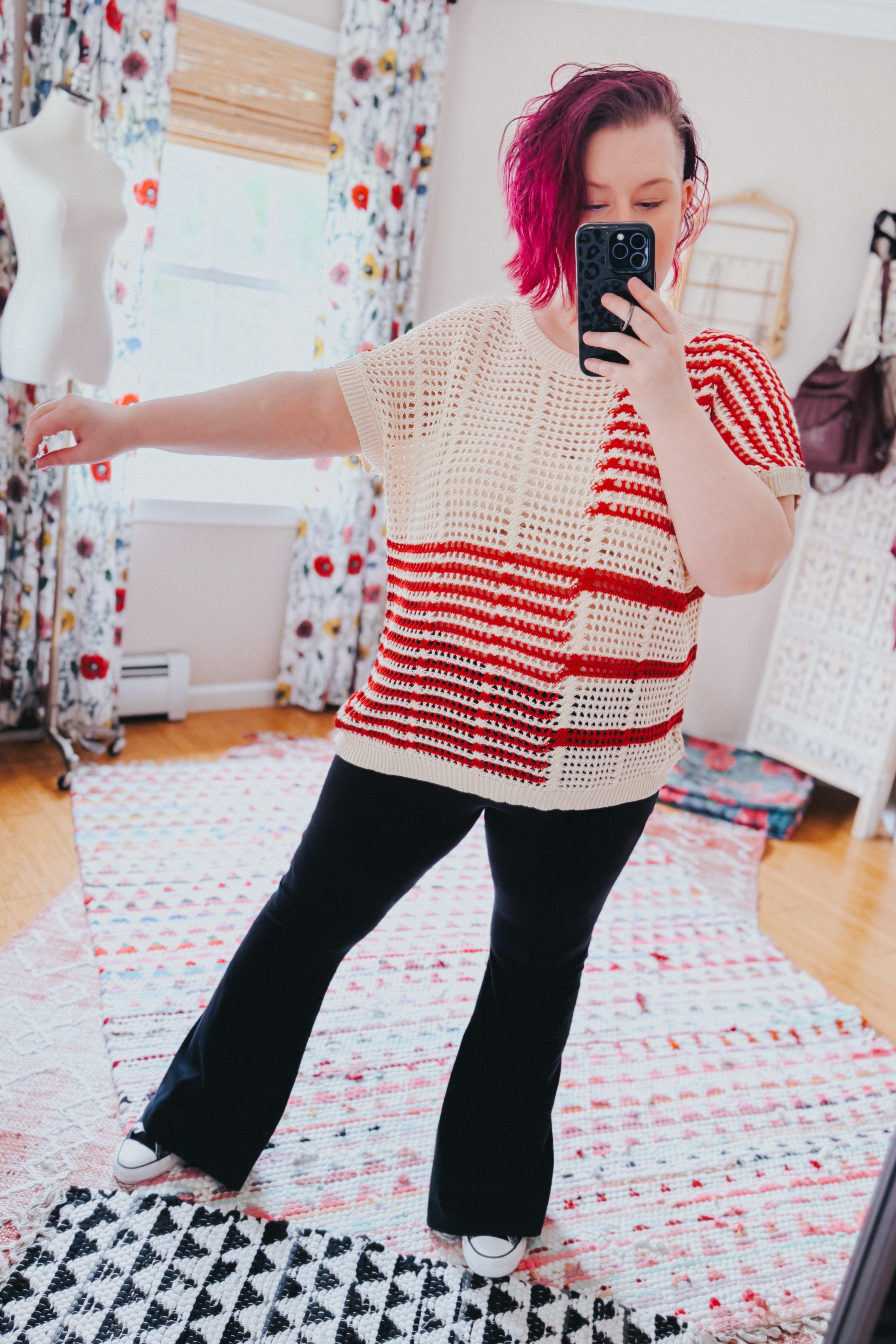 Woman with bright pink hair wearing a red striped net tee and black pants taking a mirror selfie in cozy room