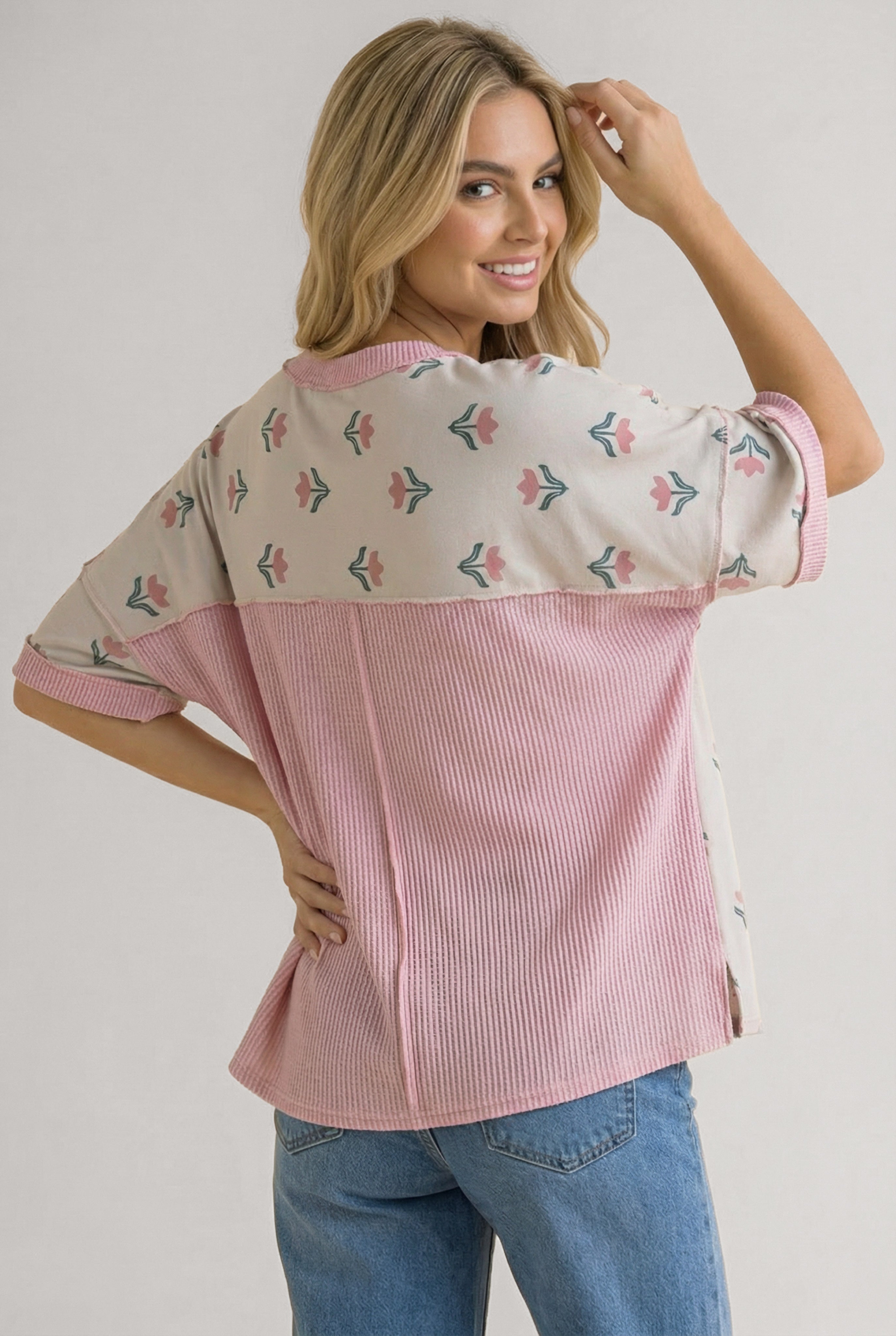 Smiling woman wearing relaxed pink floral tee with mixed fabrics and blue jeans against neutral backdrop