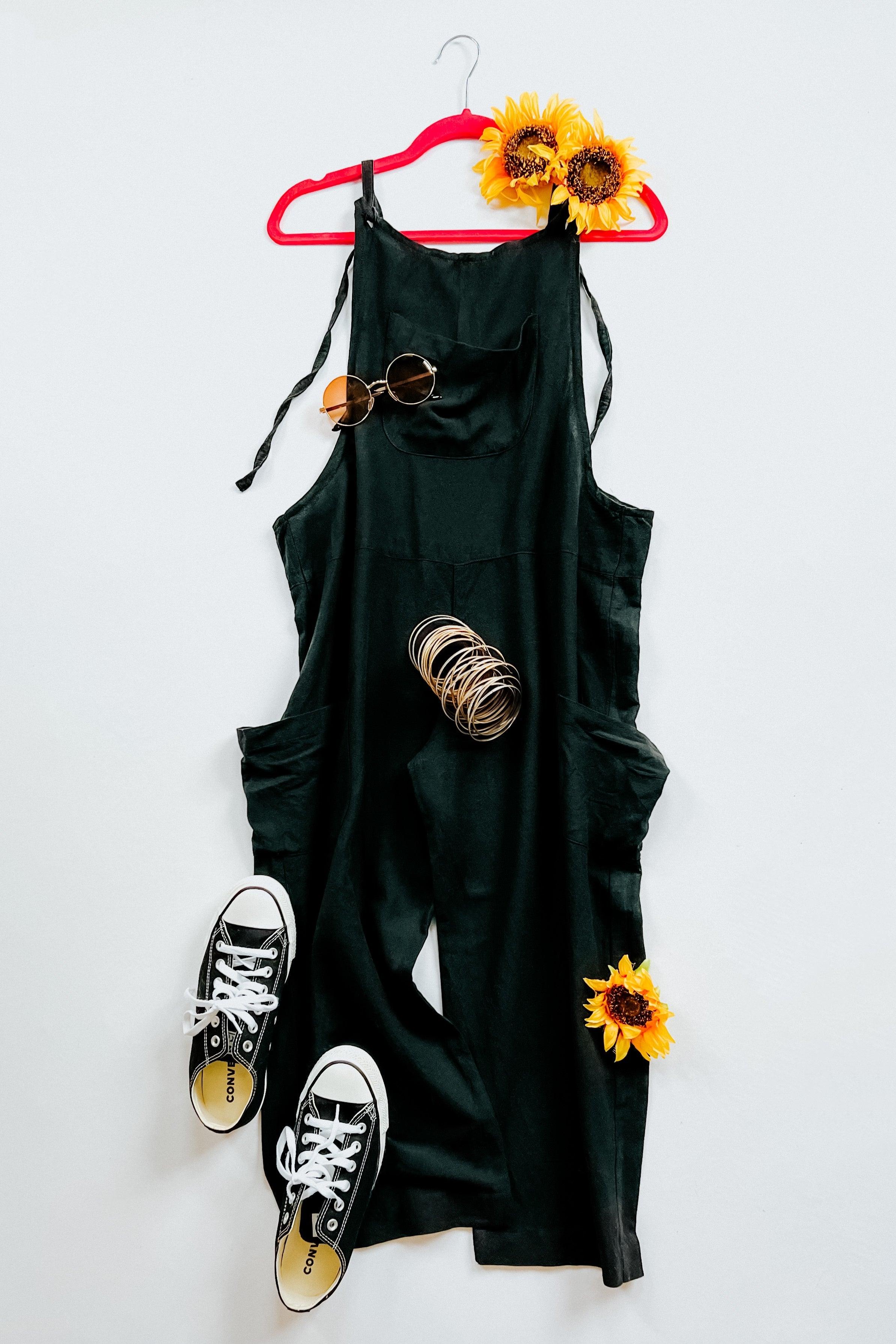 Black slouchy overalls on red hanger with sunglasses, stack of bangles, black sneakers, and sunflowers on white background
