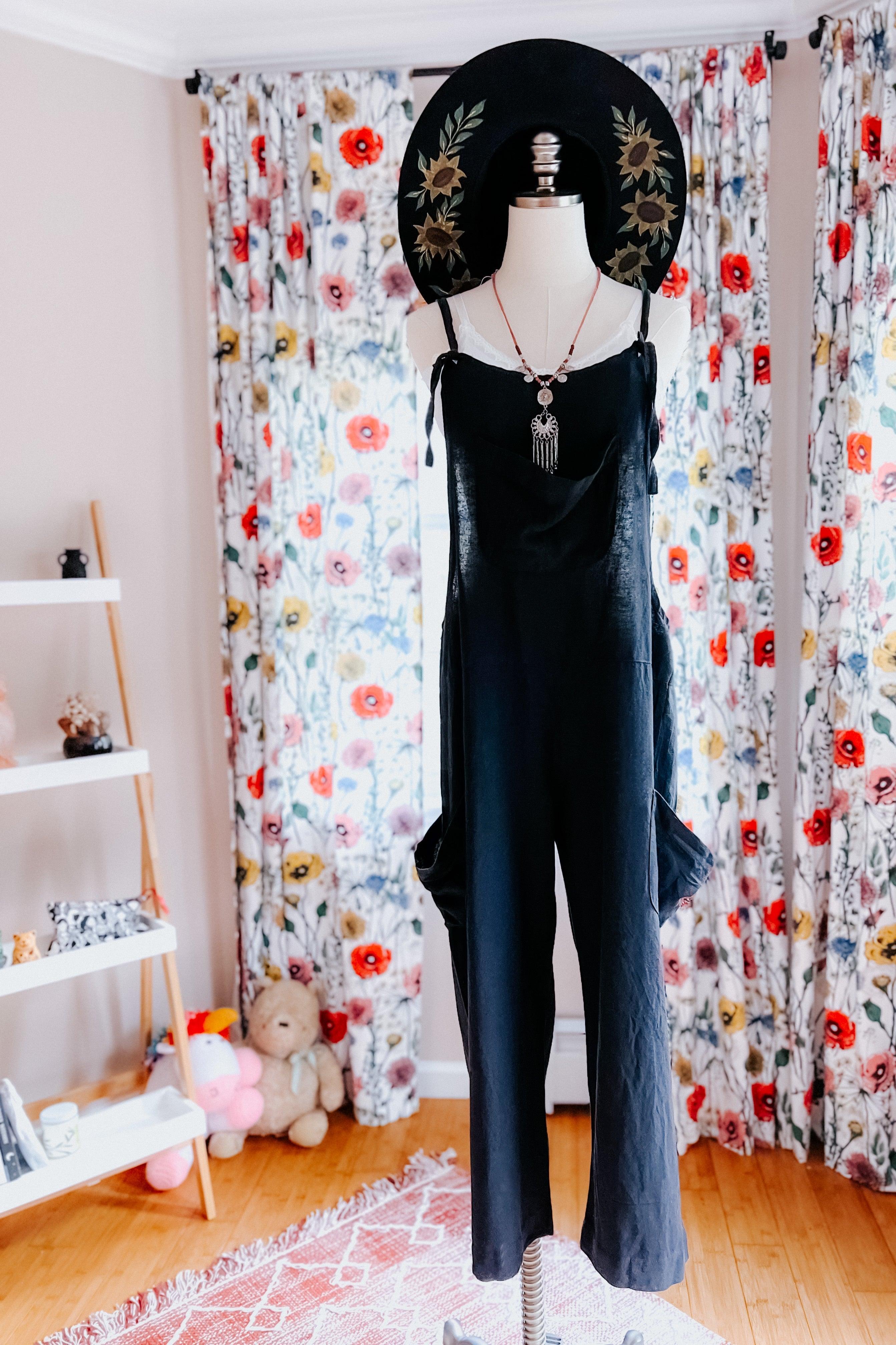 Black slouchy hemp viscose overalls styled with layered necklaces and wide-brim floral hat indoors by floral curtains