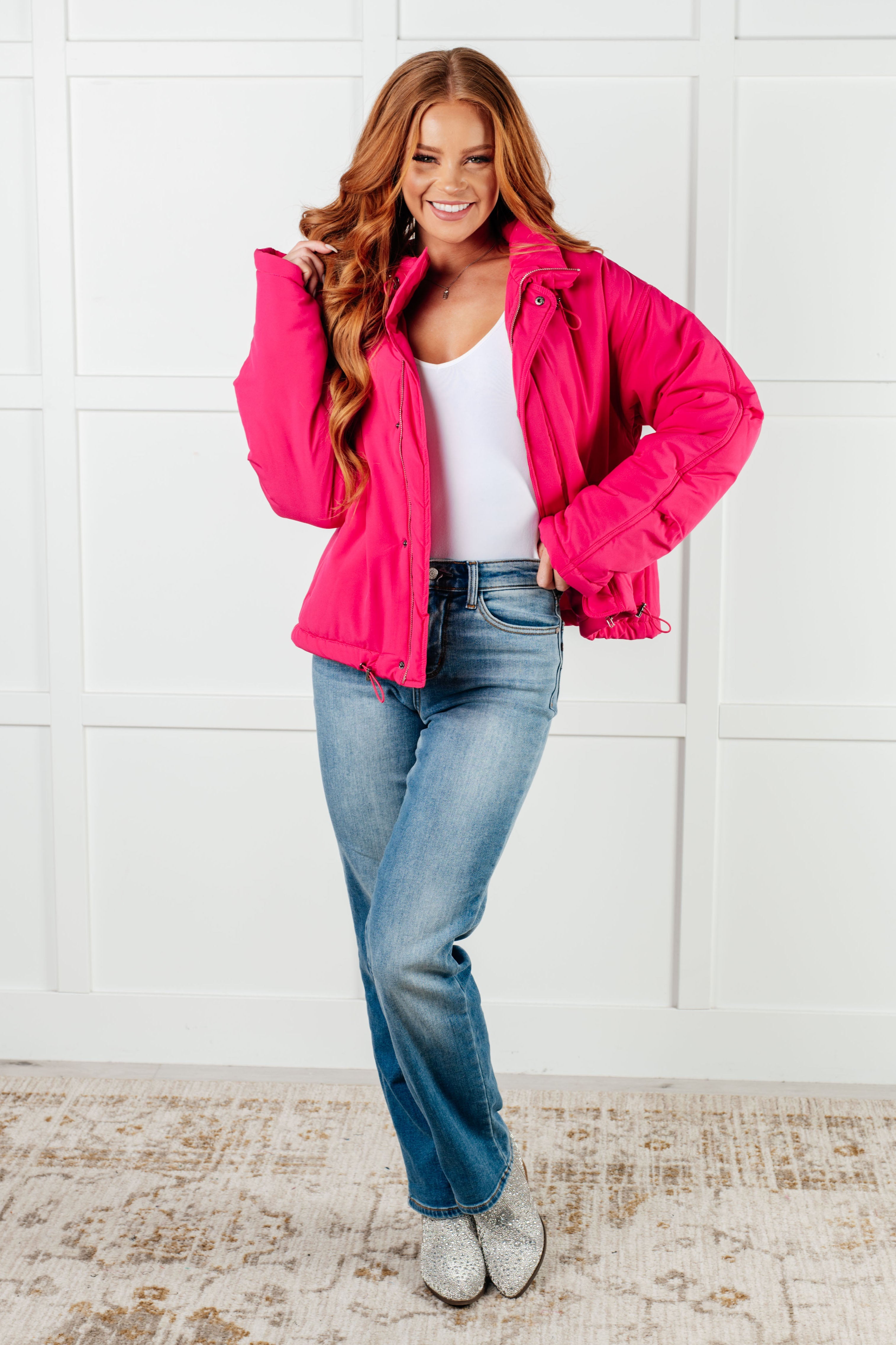 Smiling woman wearing bright pink puffer jacket over white top and jeans, posing indoors on patterned rug