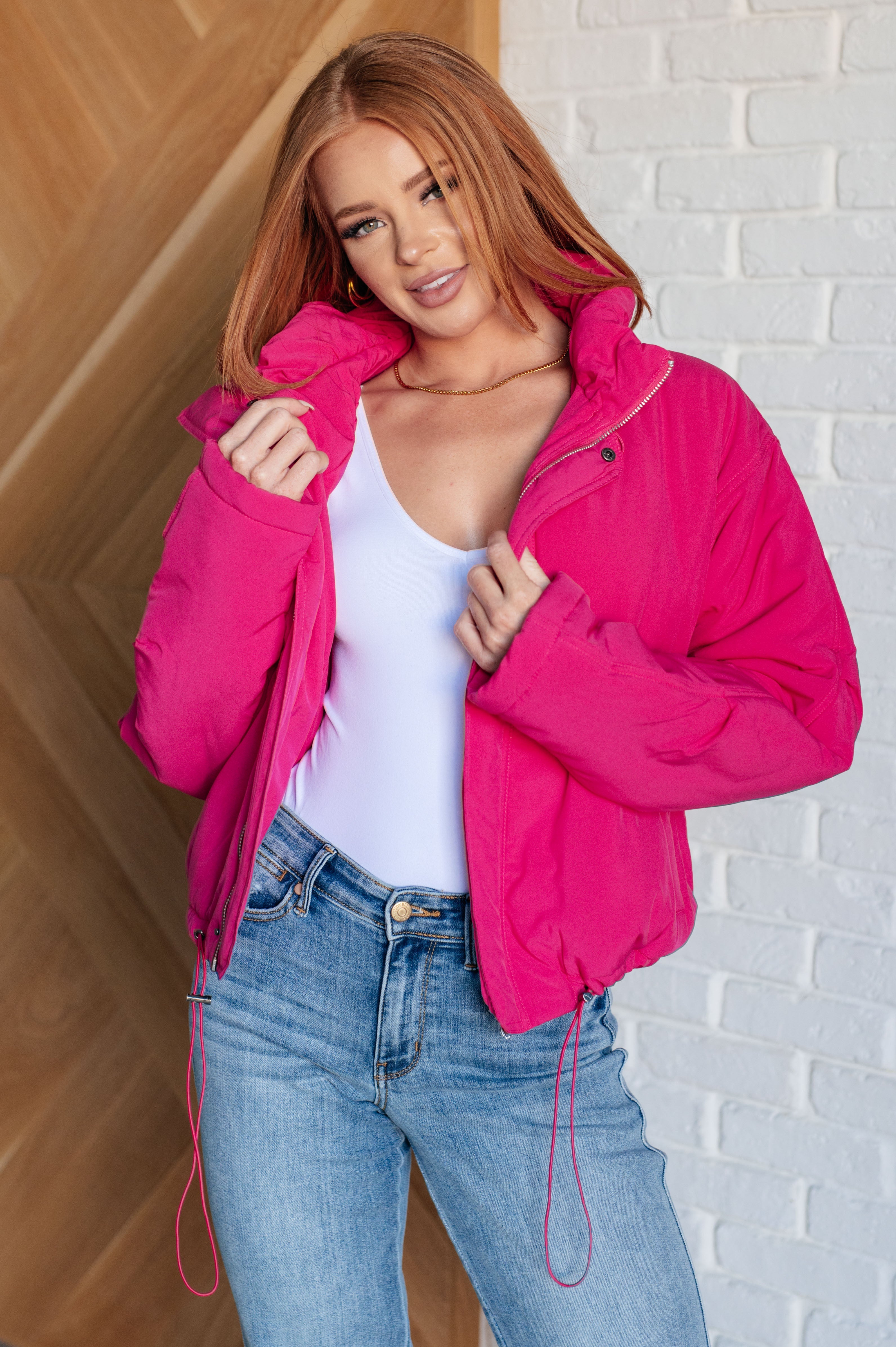 Woman wearing bright pink puffer jacket over white top and blue jeans standing indoors with relaxed expression