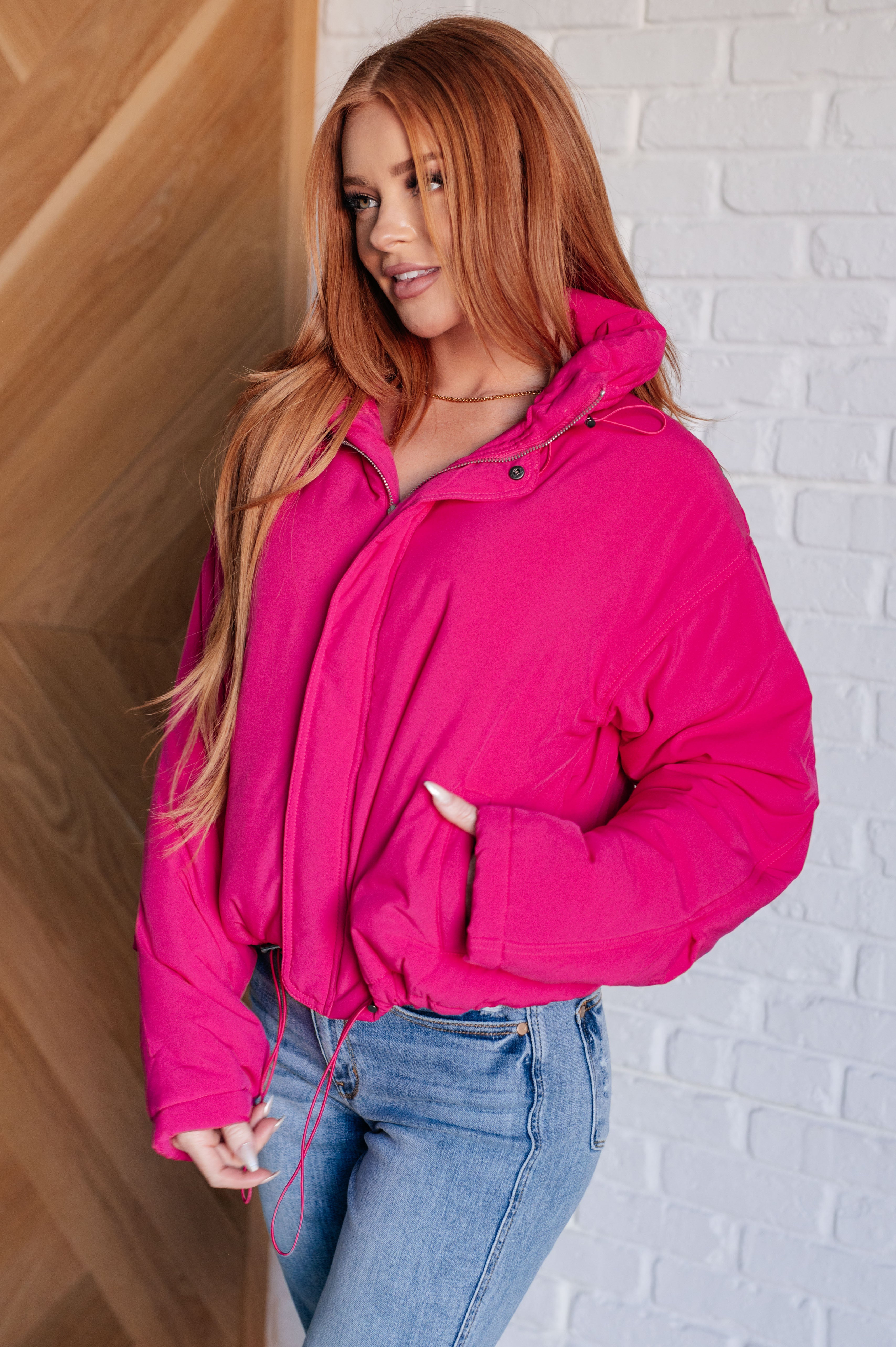 Woman with long red hair wearing vibrant pink puffer jacket styled with light wash jeans indoors
