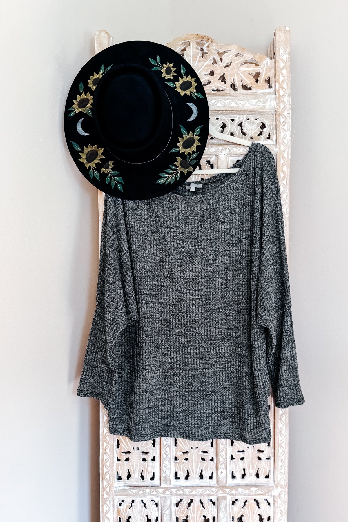 Charcoal waffle knit top with a wide boat neckline and relaxed dolman sleeves displayed on an ornate white rack beneath a black floral hat