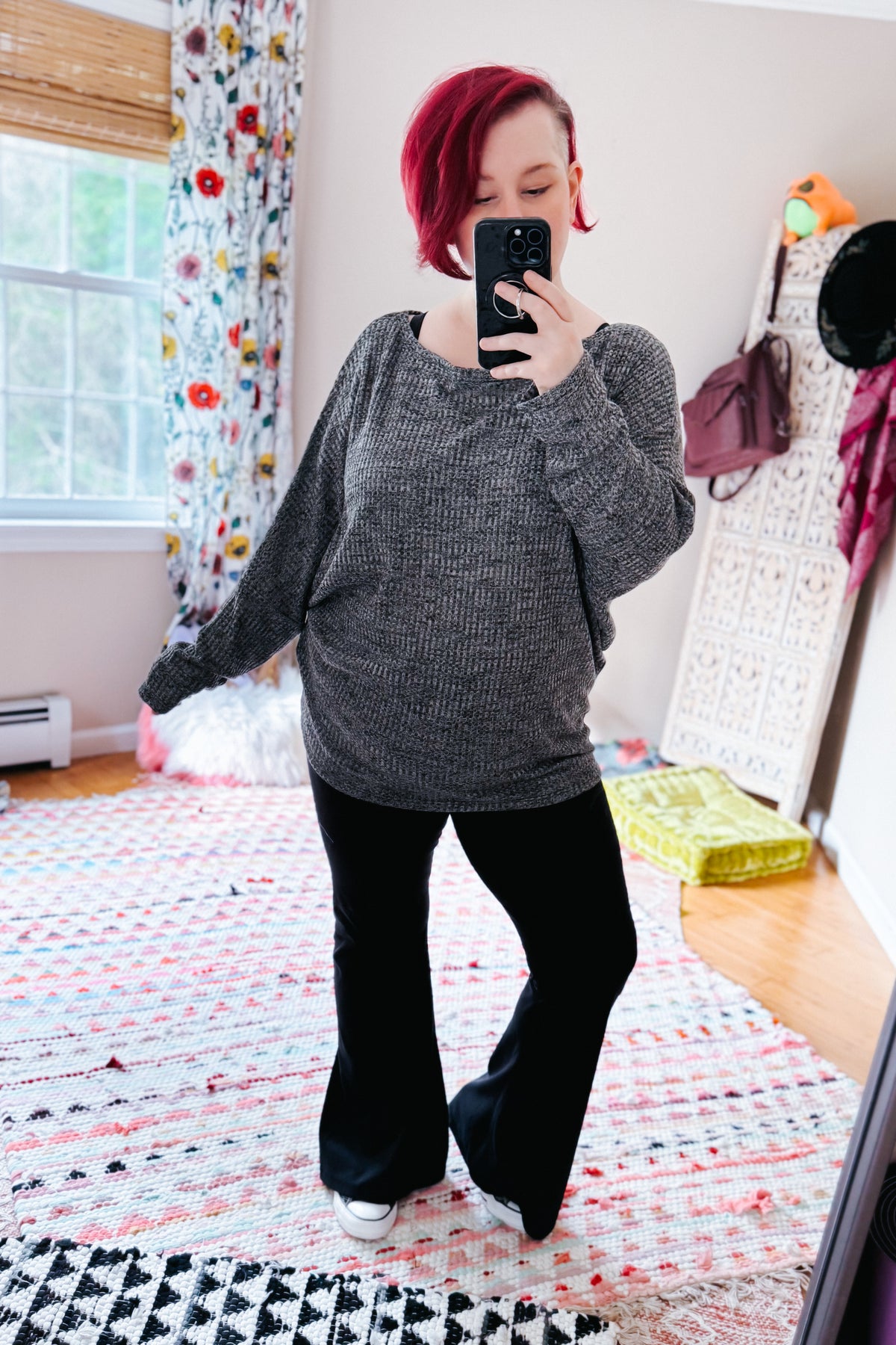 Red haired woman wearing a charcoal ribbed dolman sleeve top with black flared pants and white sneakers in a colorful bedroom mirror selfie