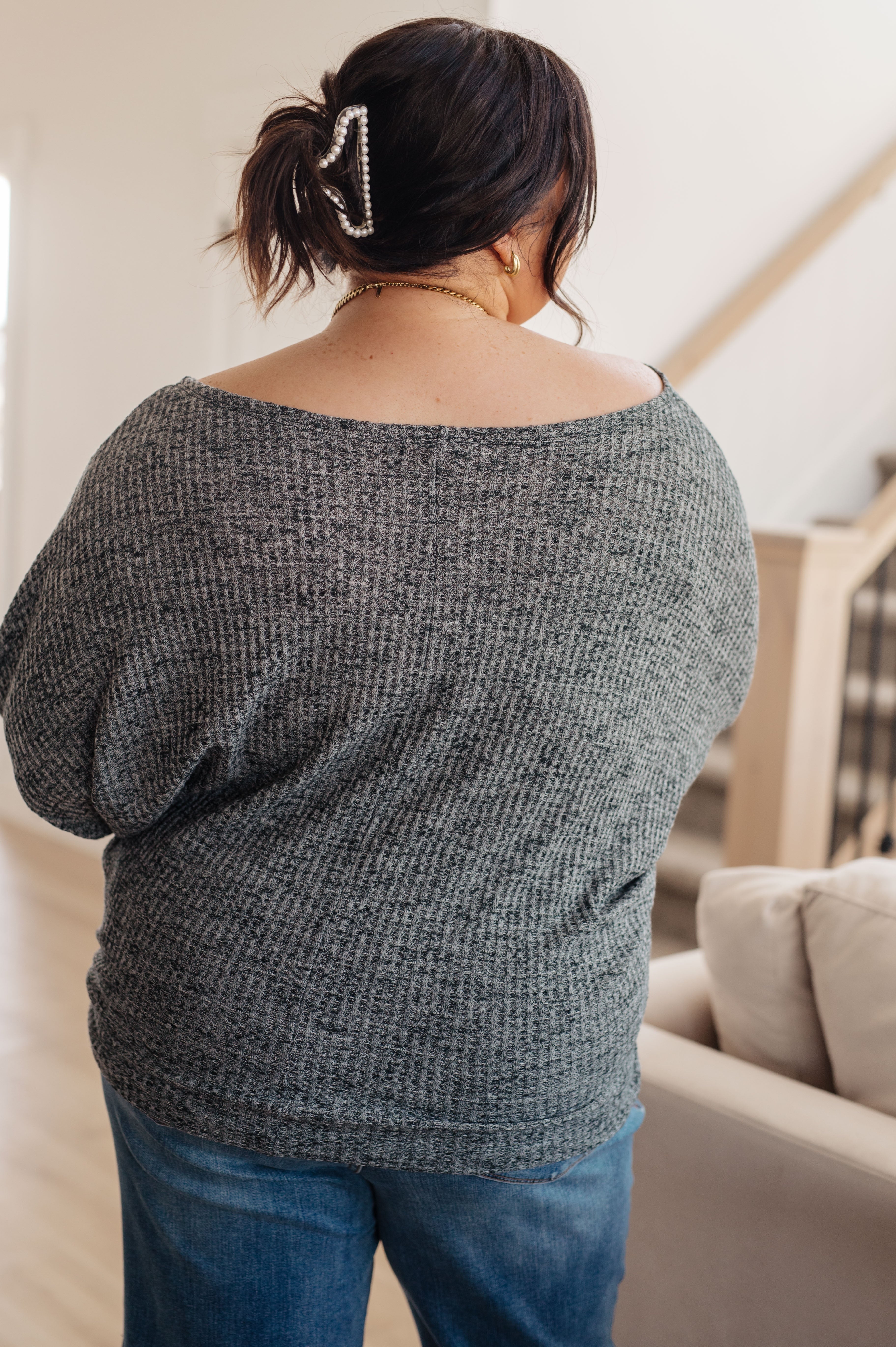 Woman wearing charcoal ribbed top with off-shoulder design and blue jeans indoors by beige couch and stairs