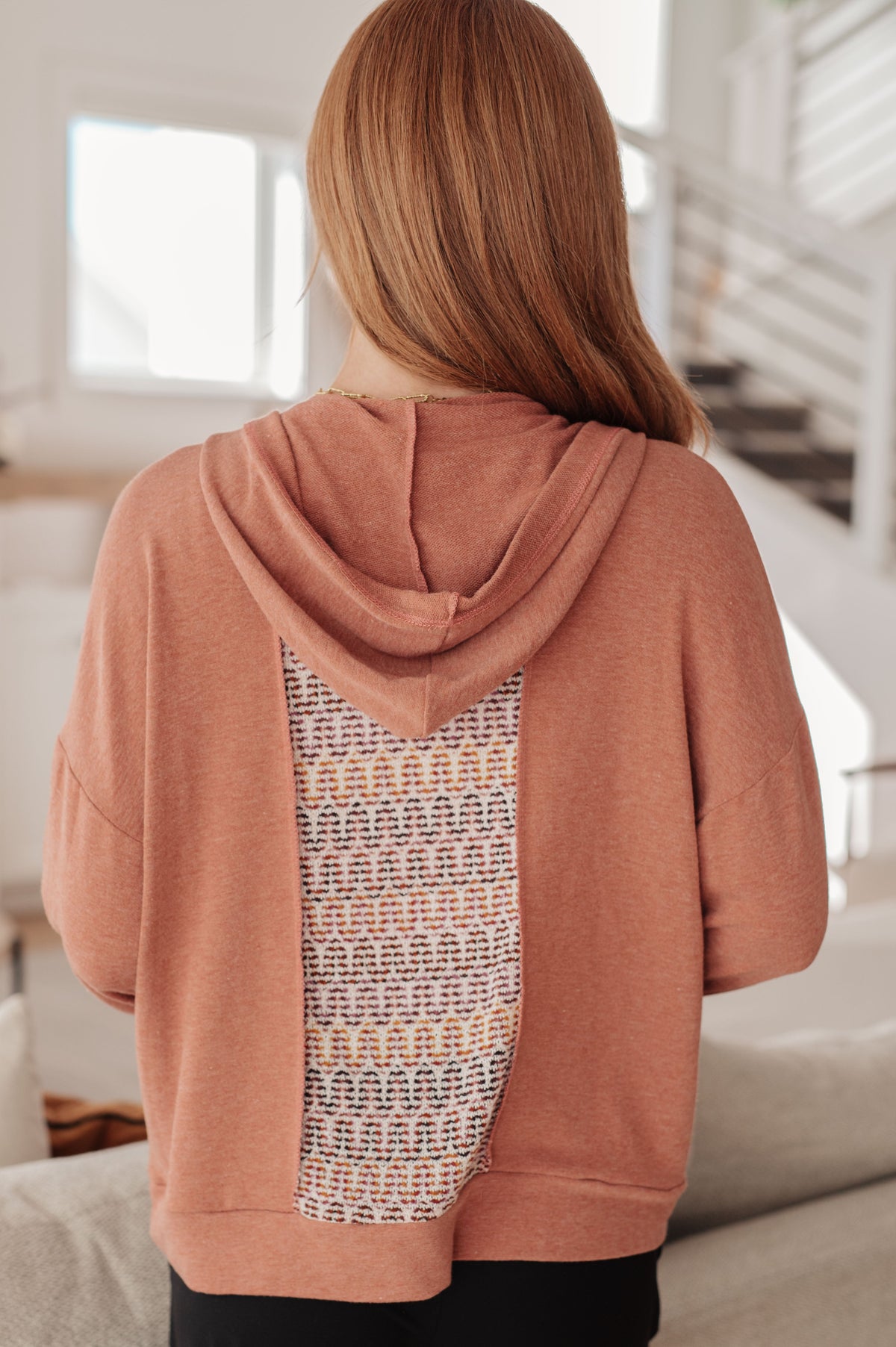 Back view of a woman wearing a rust hooded top with a multicolored textured back panel and black pants indoors