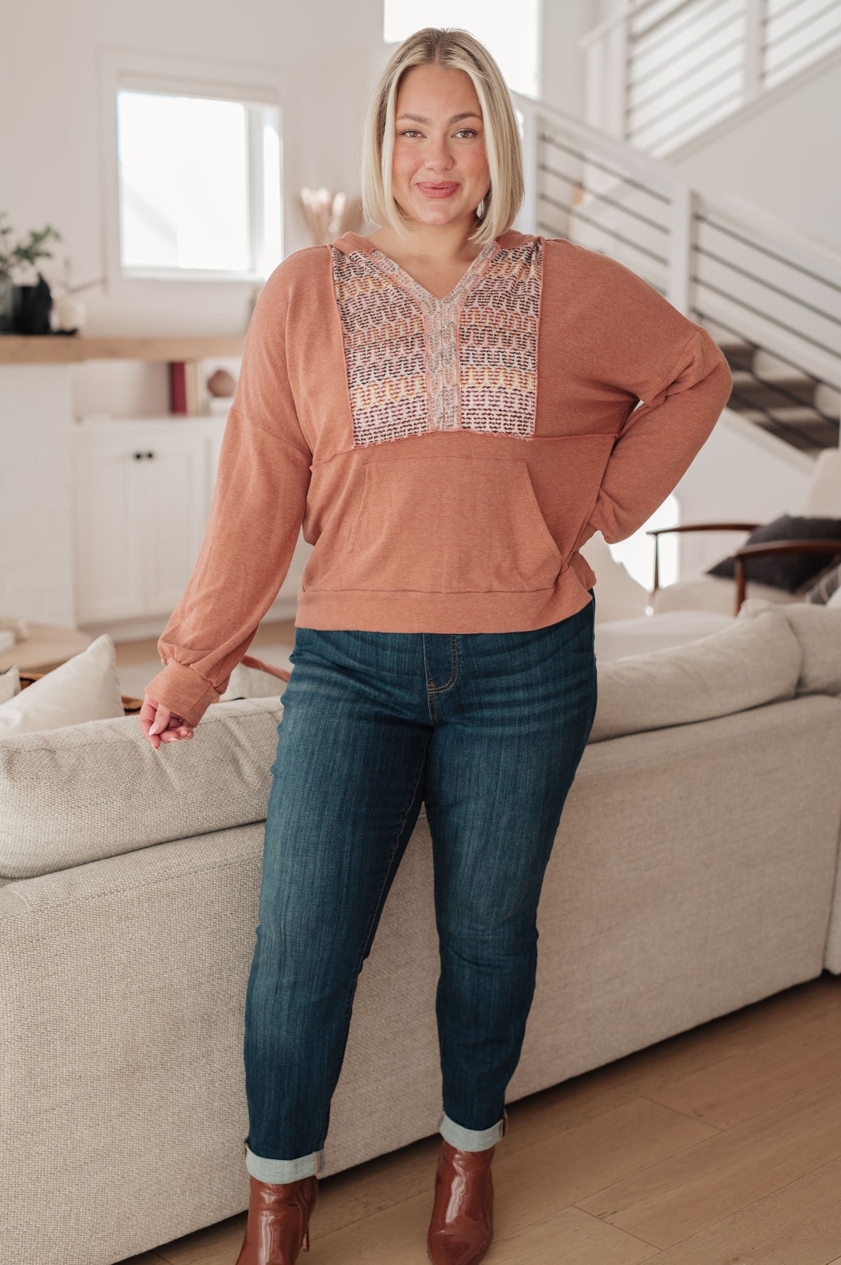Woman modeling a rust hooded jersey top with a multicolored textured front panel and kangaroo pocket with cuffed jeans and boots indoors
