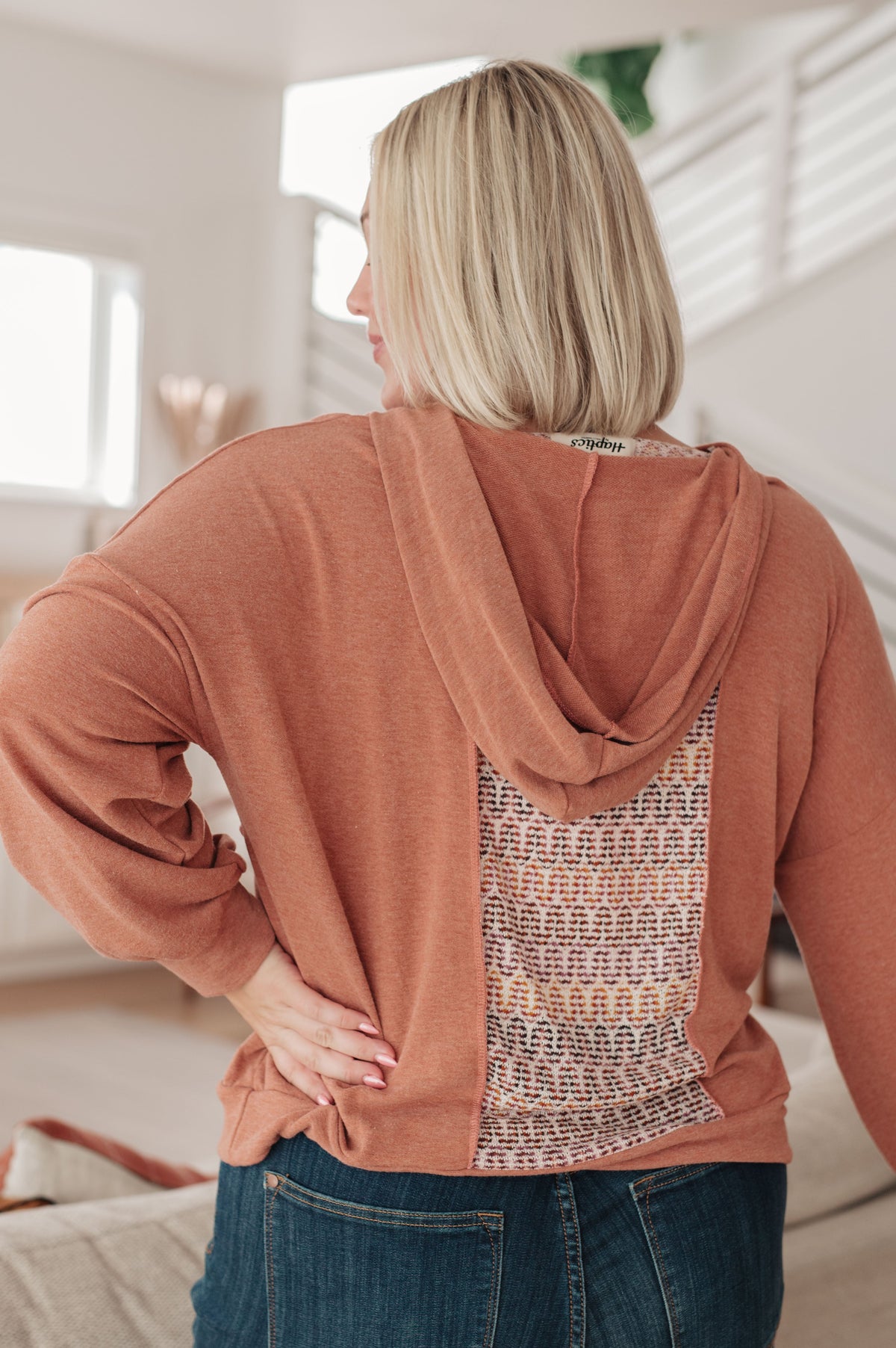 Back view of a woman wearing a rust hooded top with a multicolored textured back panel and jeans in a bright living room