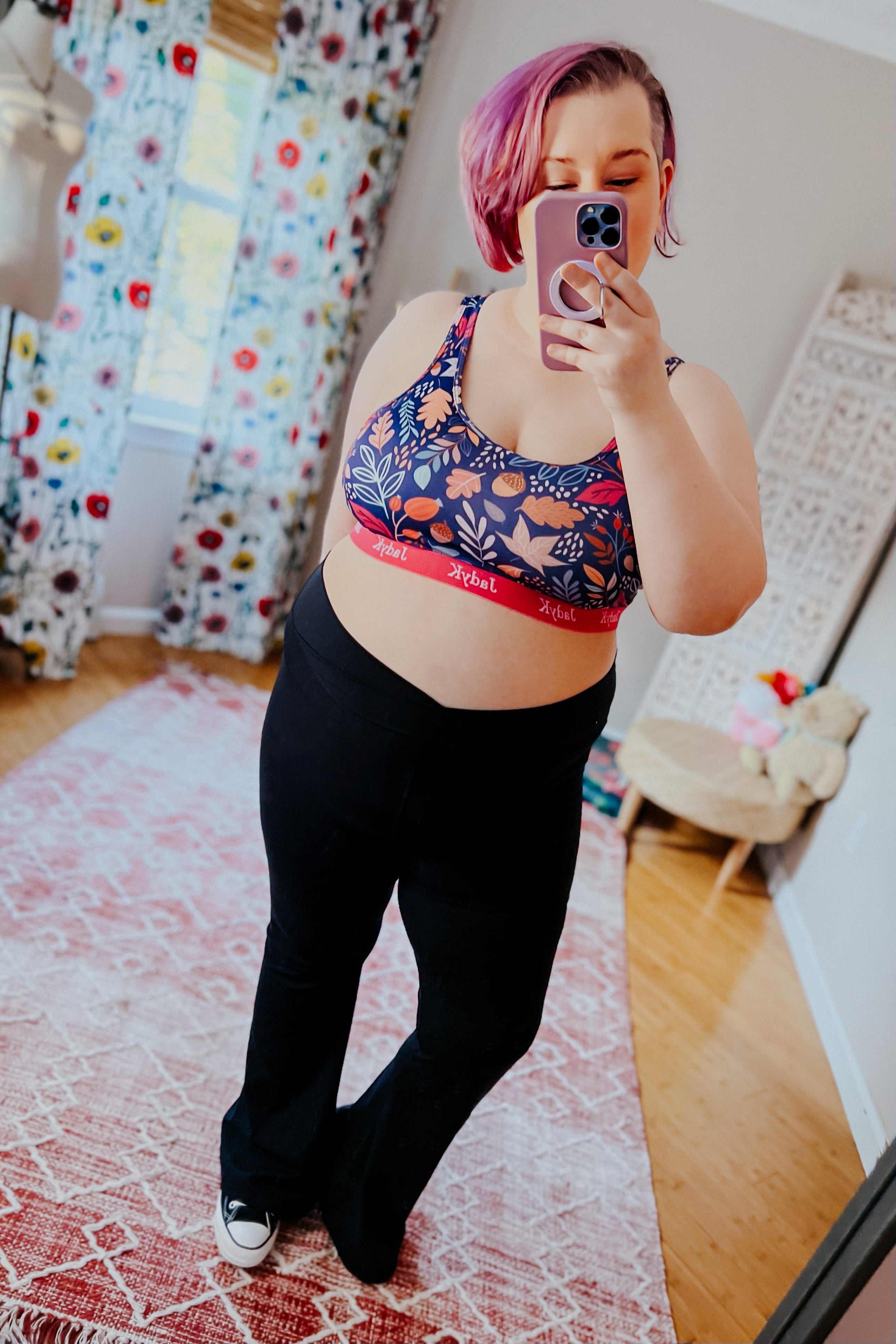Woman with purple hair wearing blue patterned bra with red band and black pants taking a mirror selfie in cozy room