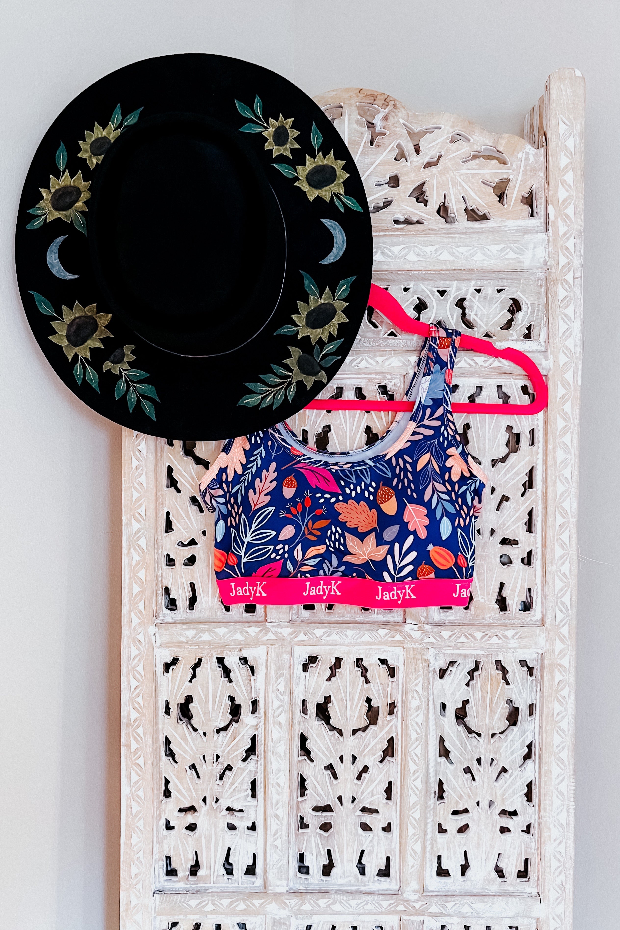 Blue scoop bra with floral and leaf print and pink band hanging on carved white wooden panel next to black hat with sunflower design