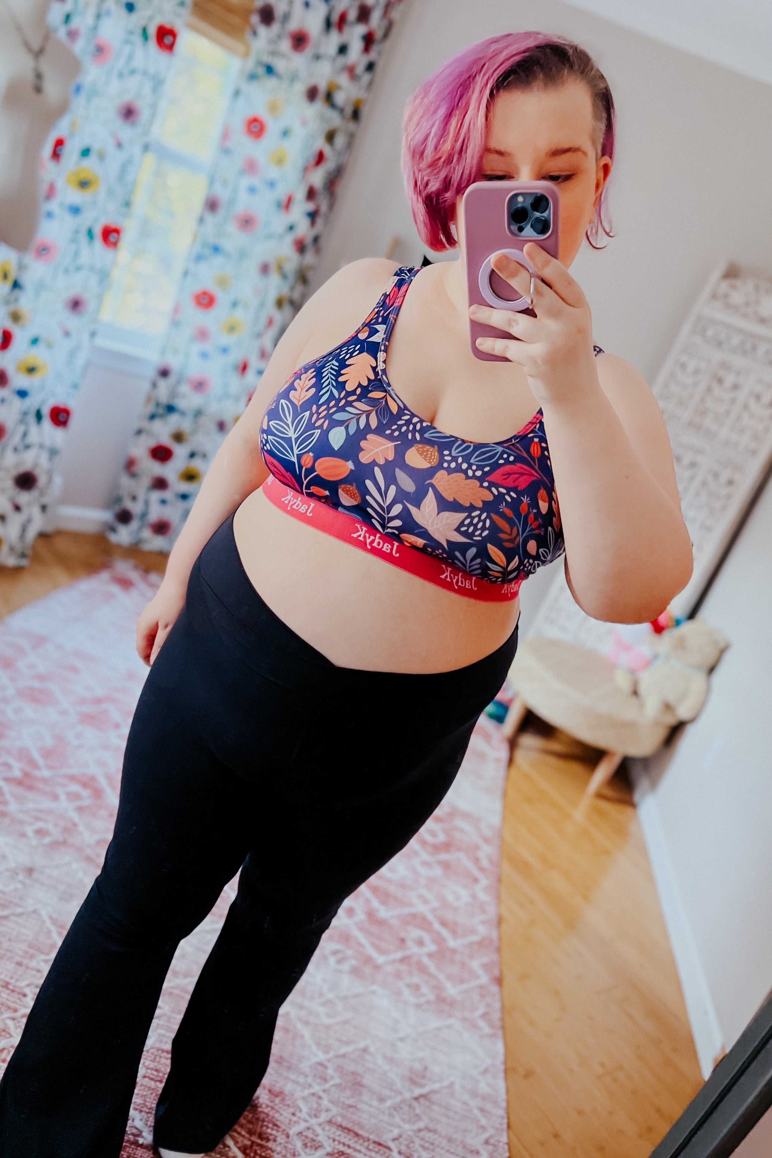 Plus-size woman with short pink hair wearing blue floral patterned sports bra and black leggings taking mirror selfie indoors