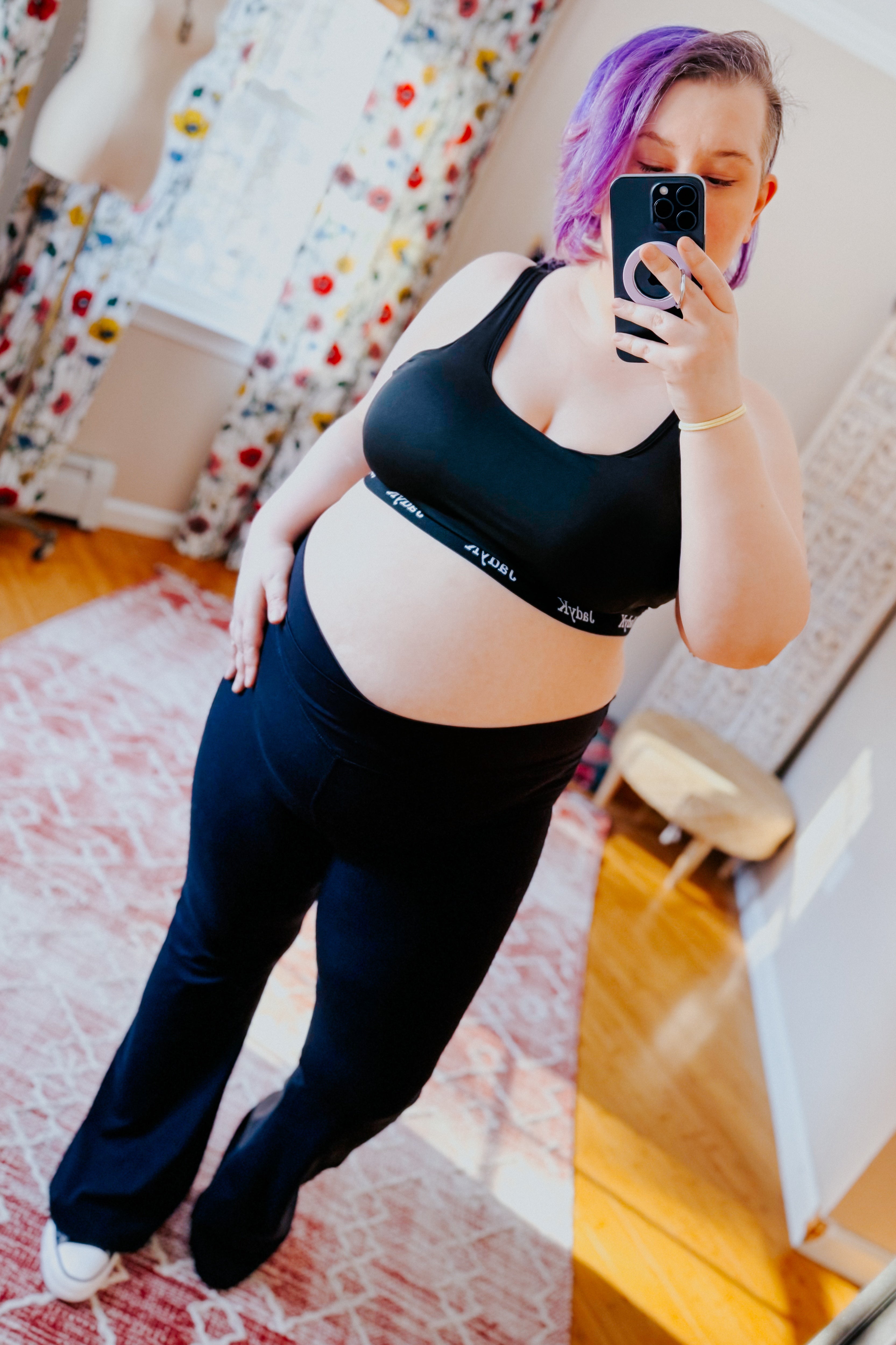 Woman with purple hair wearing black scoop bralette and pants taking a cozy mirror selfie in sunflower-patterned room
