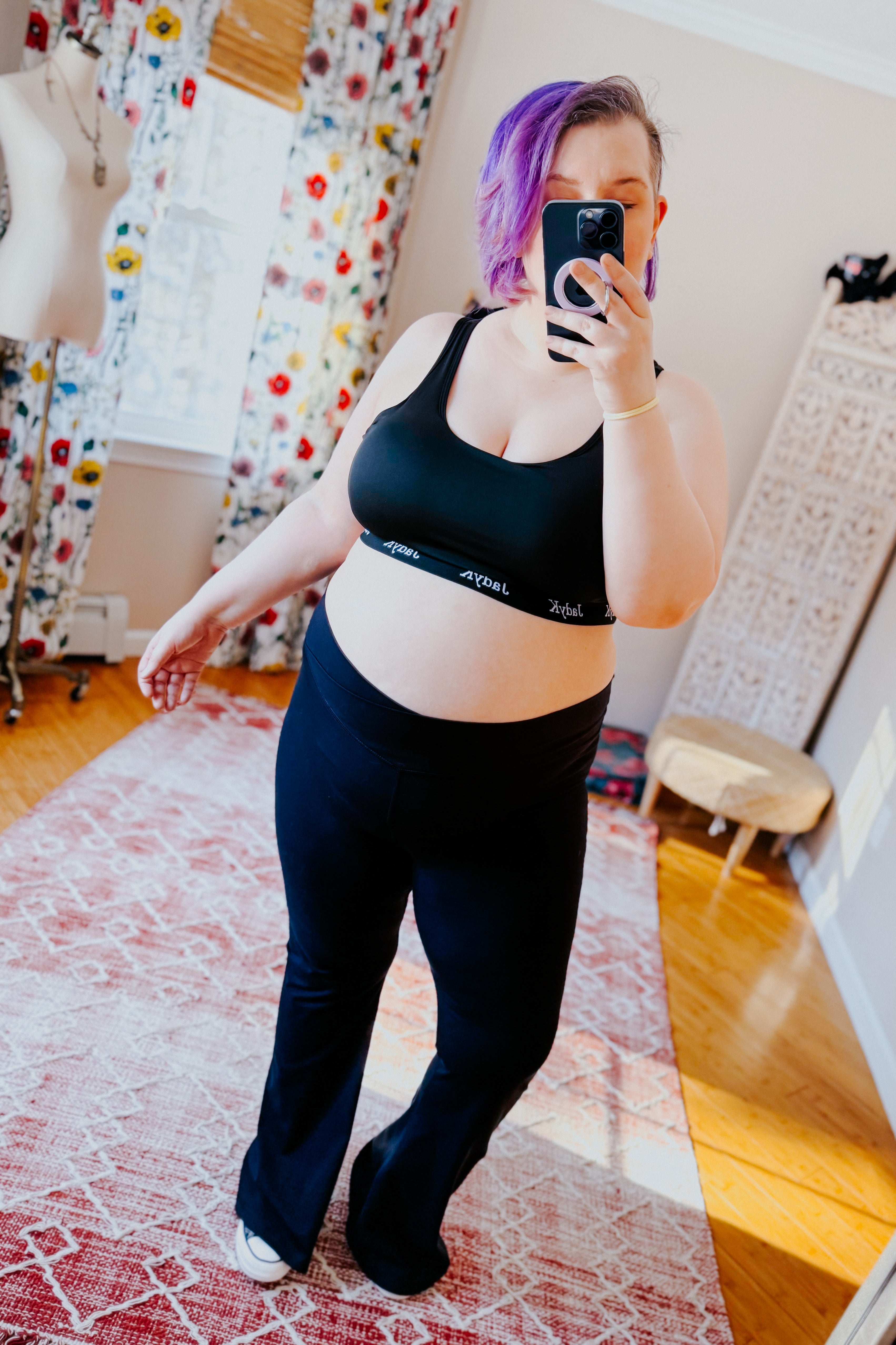 Plus-size woman with purple hair wearing black bralette and matching pants taking a mirror selfie in bright room