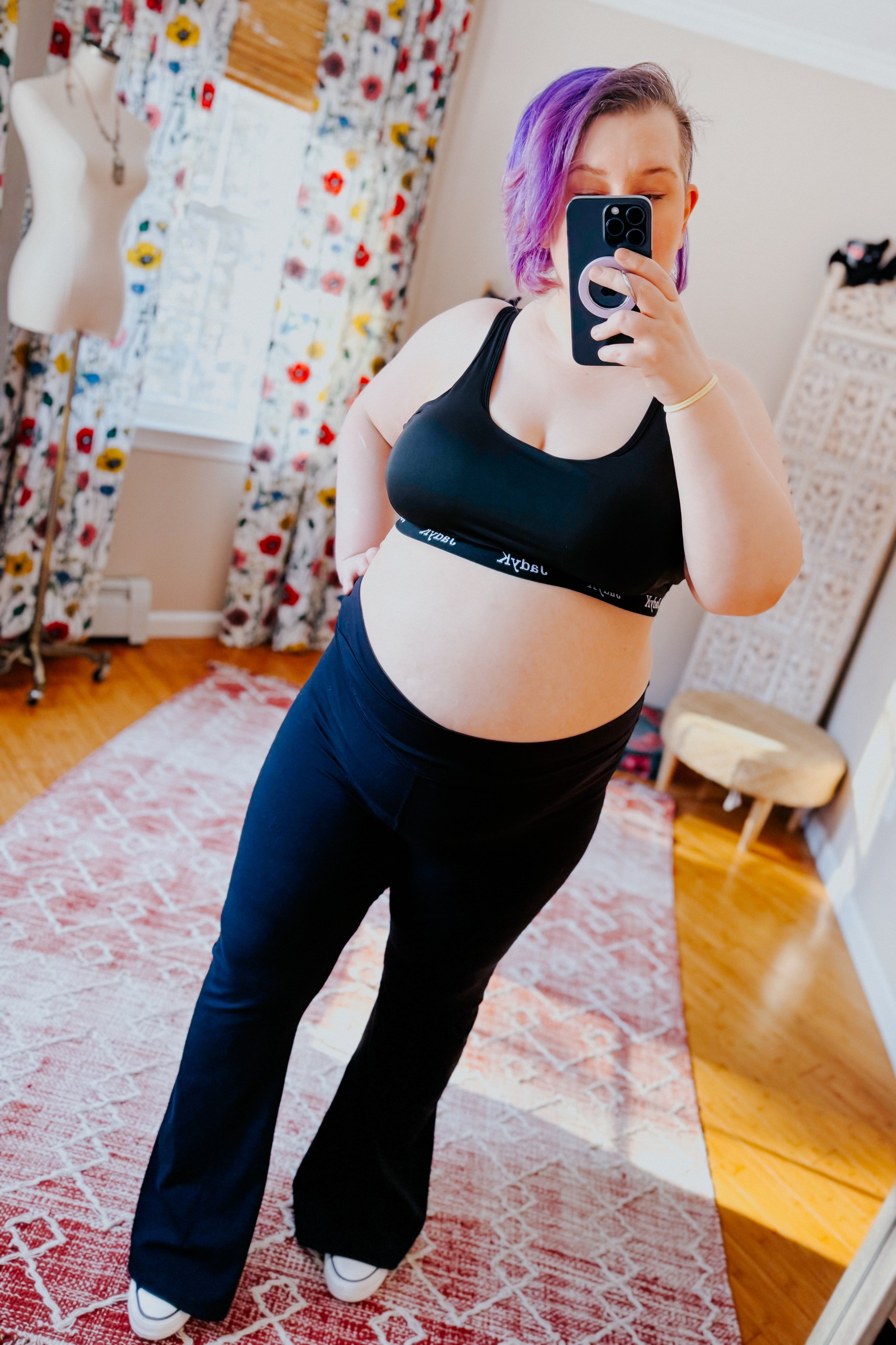 Plus-size woman with purple hair wearing form-fitting black bralette and pants taking a mirror selfie in bright room