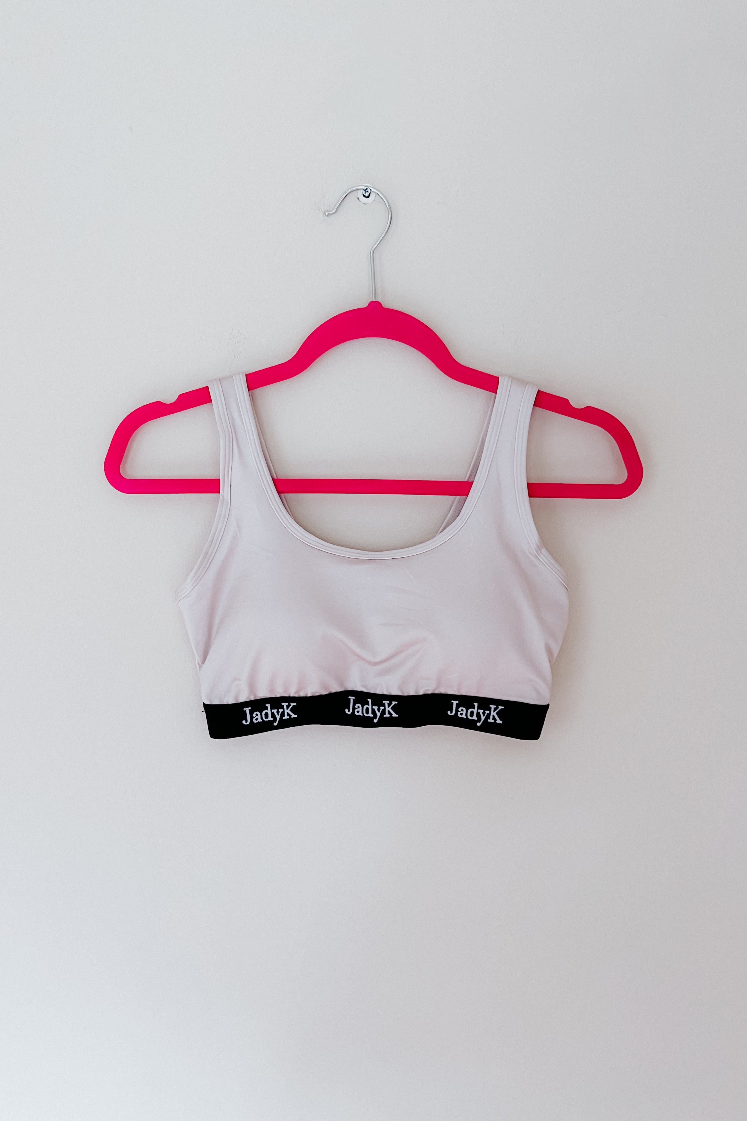 Light champagne bralette with branded black elastic band hanging on vibrant pink hanger against white wall background