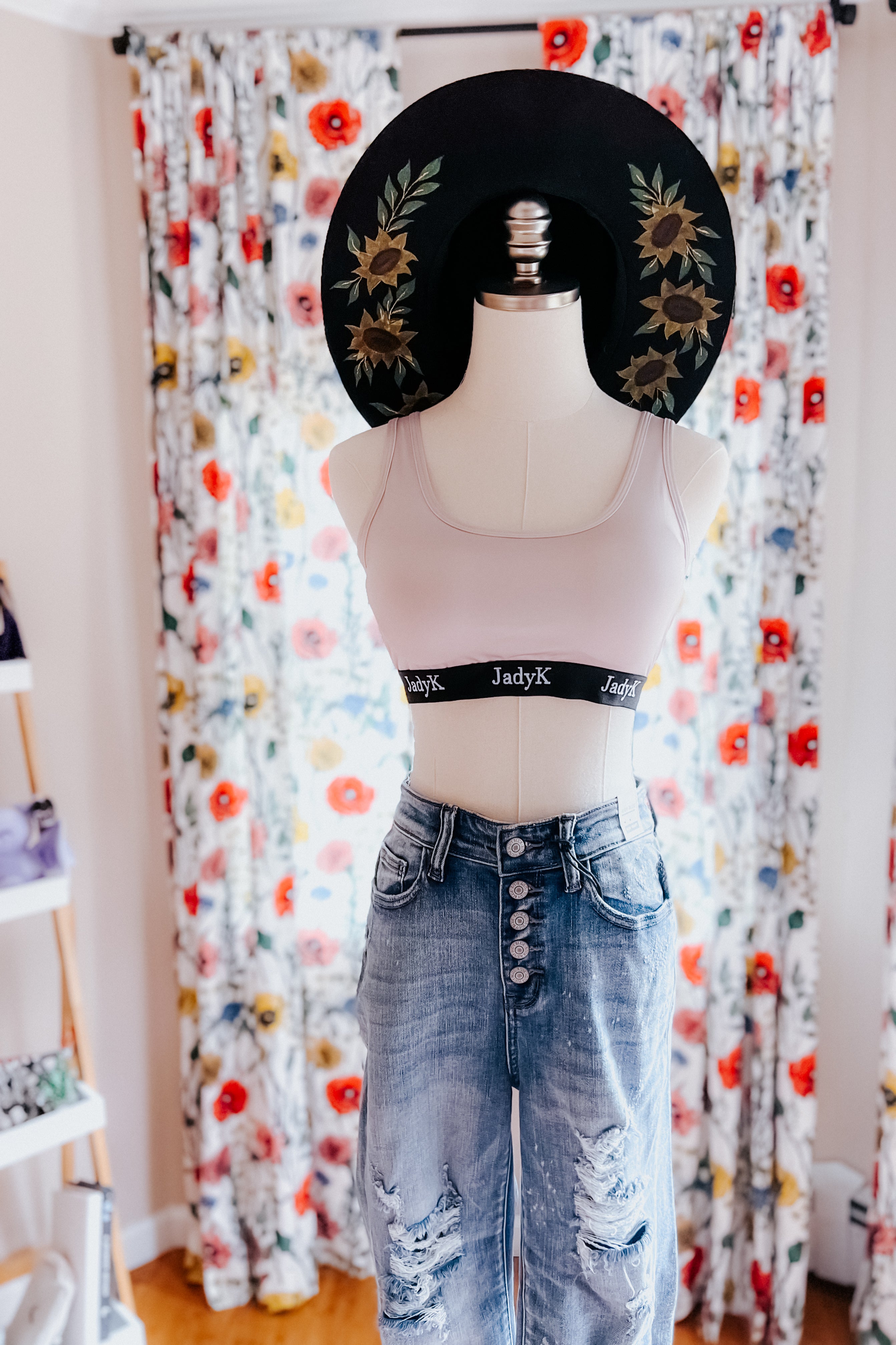 Mannequin dressed in champagne bralette with logo band paired with distressed high-waist jeans and black floral hat
