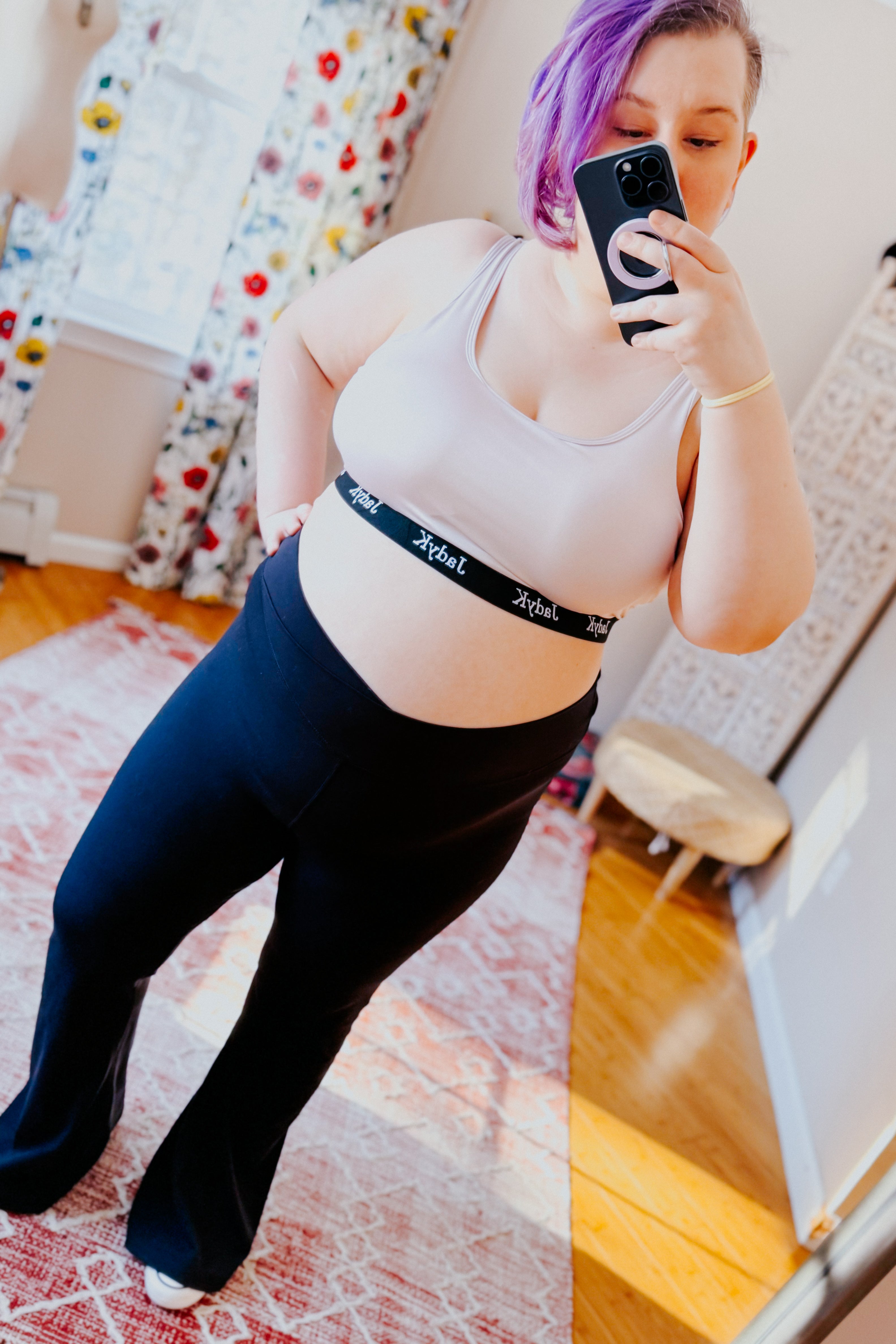 Woman with purple hair wearing champagne bralette and black pants taking a mirror selfie in a bright room with floral curtains