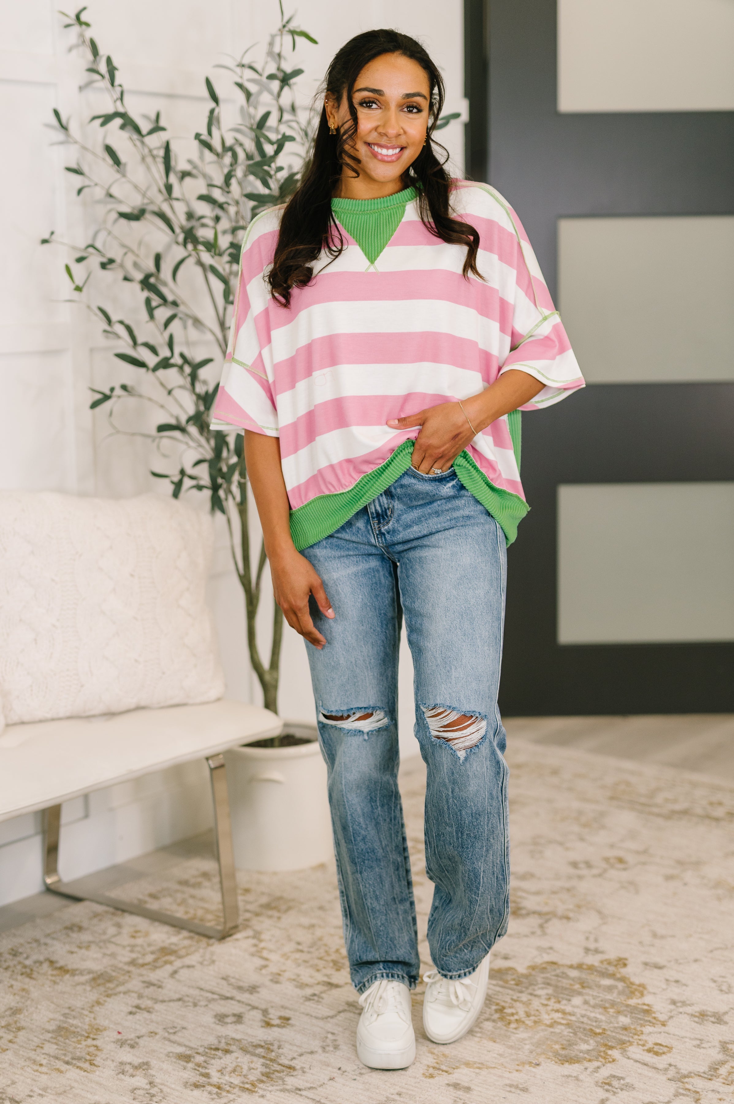 Woman wearing relaxed pink and white striped top with green trim paired with ripped blue jeans and white sneakers indoors