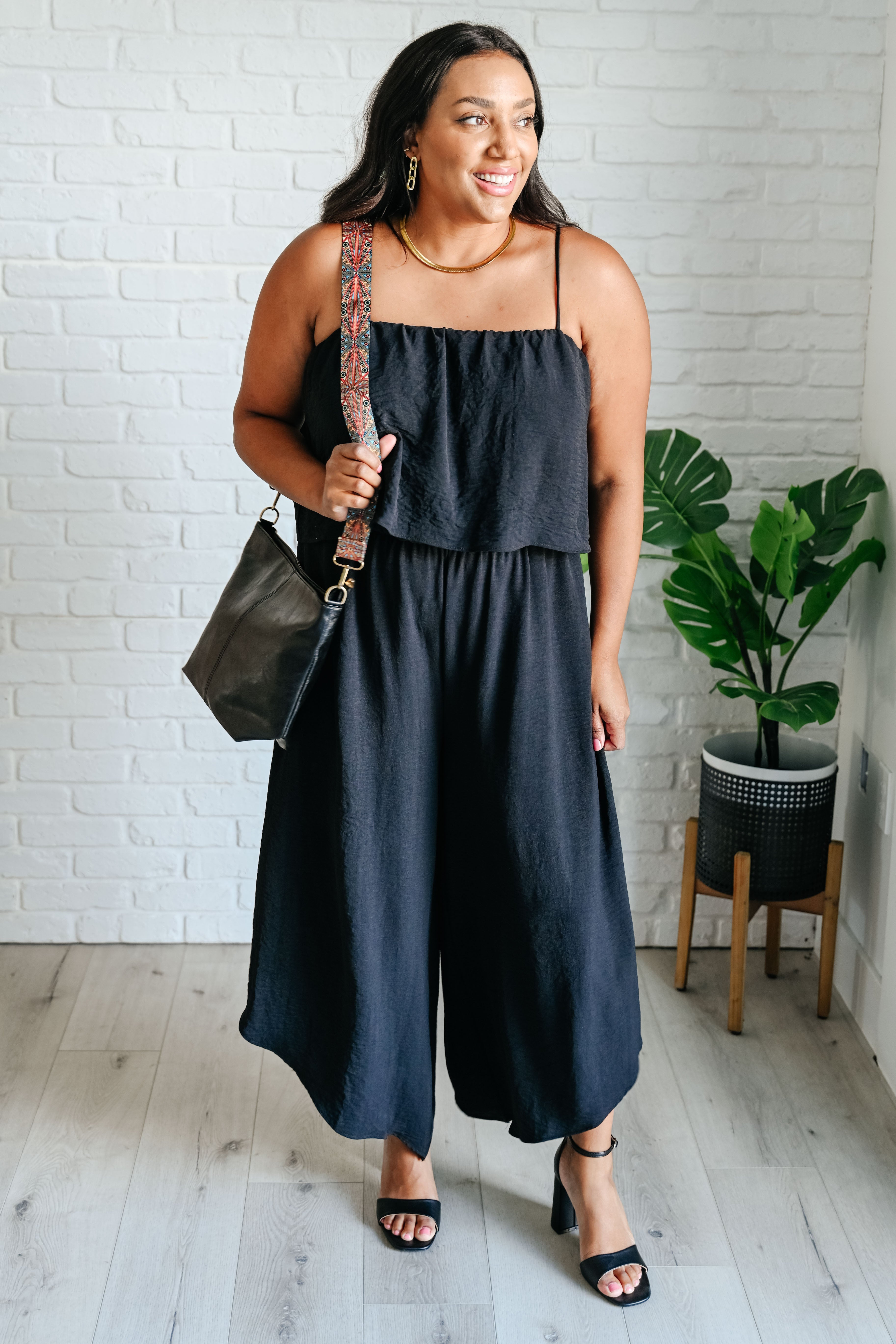 Woman wearing a black sleeveless wide-leg jumpsuit with a patterned shoulder bag and black open-toe heels indoors