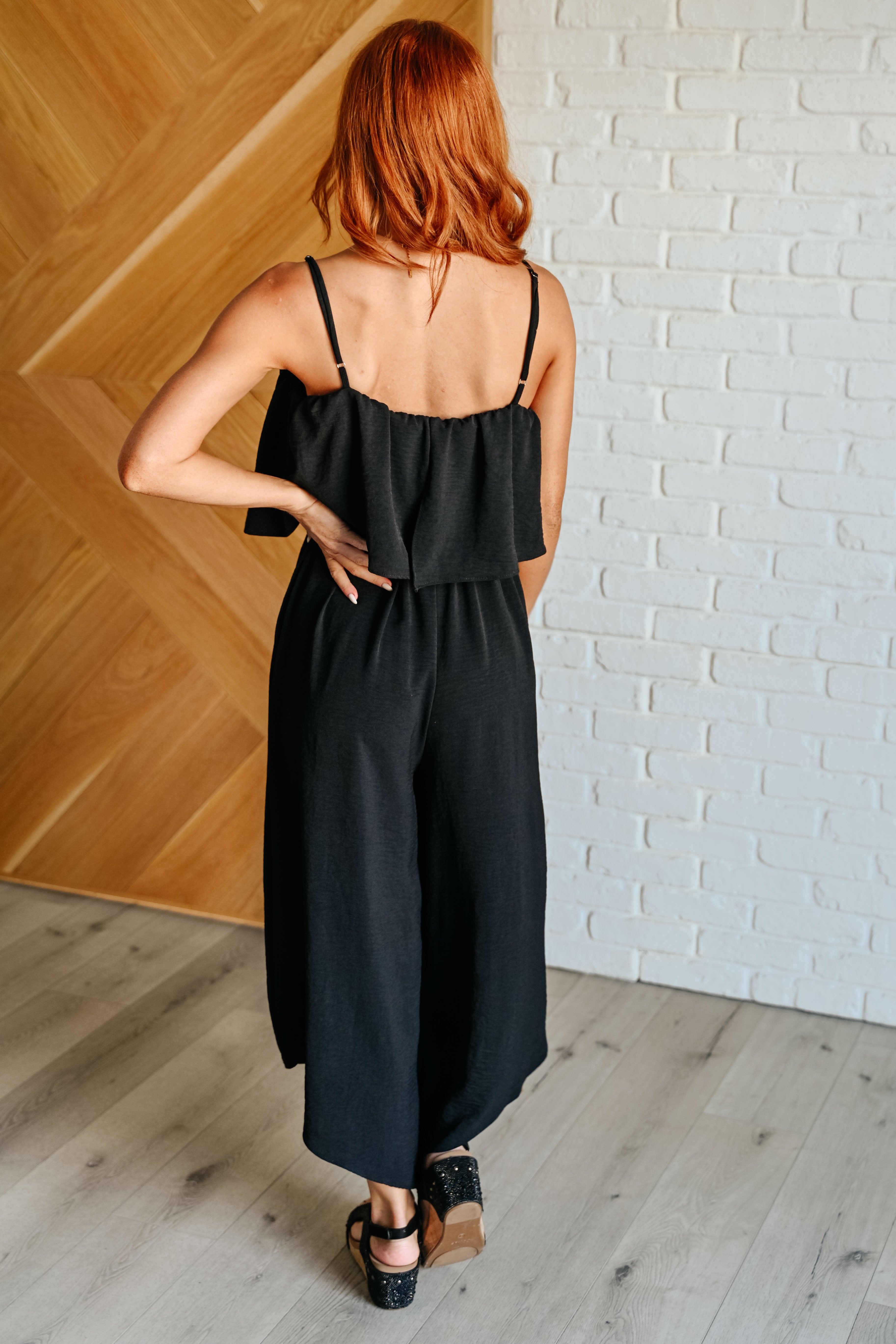 Woman with red hair wearing a black sleeveless jumpsuit with wide legs and open-back detailing indoors