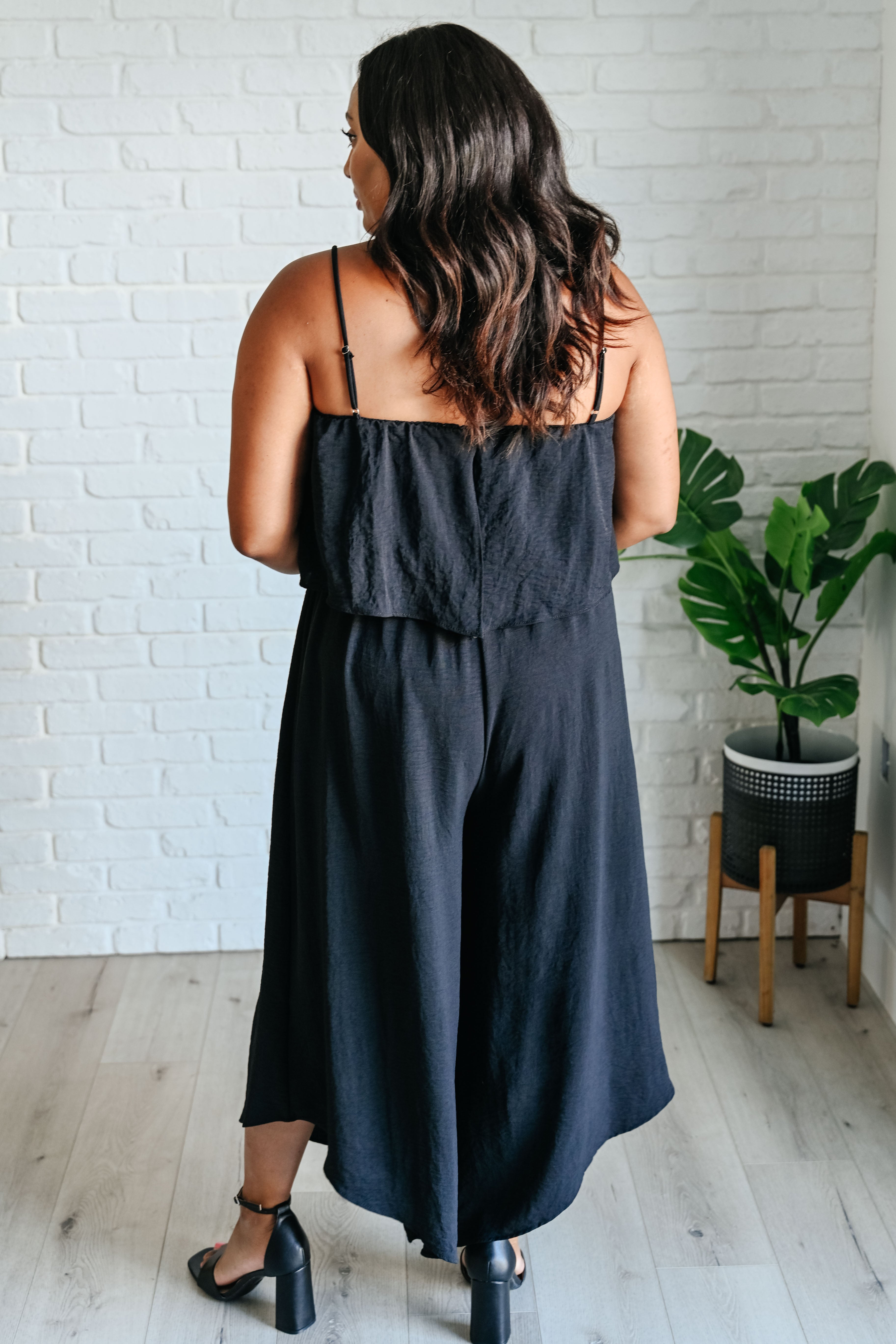 Rear view of woman wearing a black sleeveless jumpsuit with adjustable thin straps and wide-leg cropped pants indoors