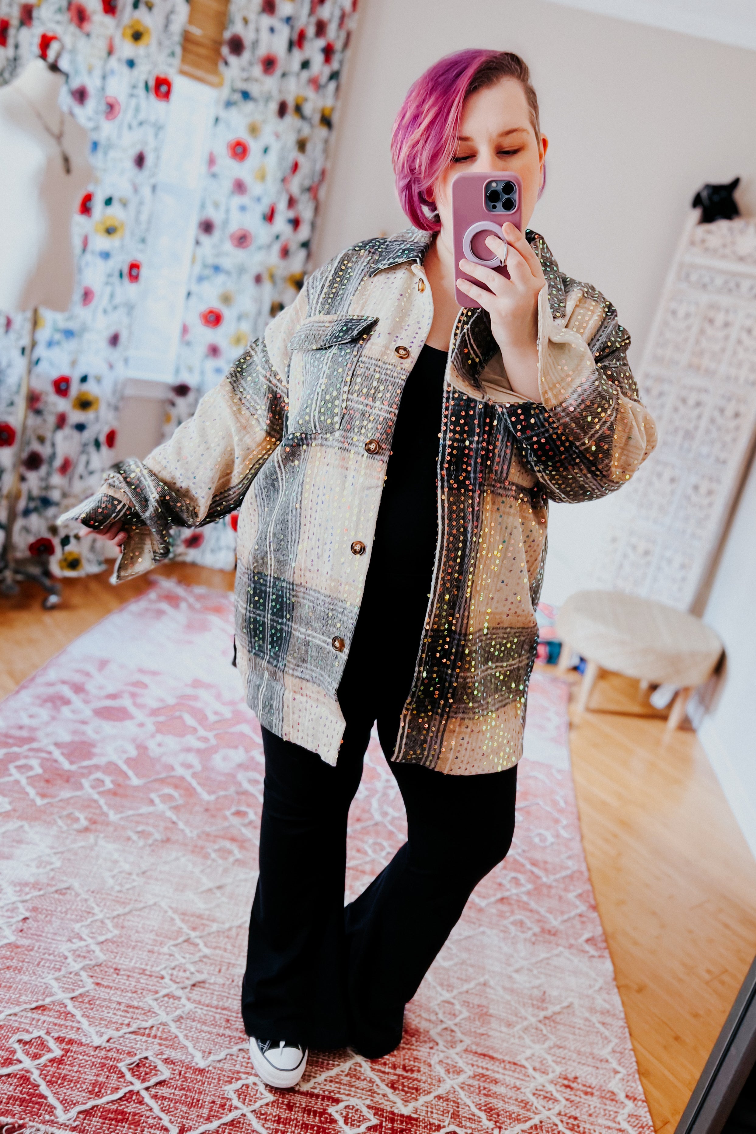 Woman with pink hair wearing beige and black plaid sequin shacket and black outfit taking a mirror selfie indoors