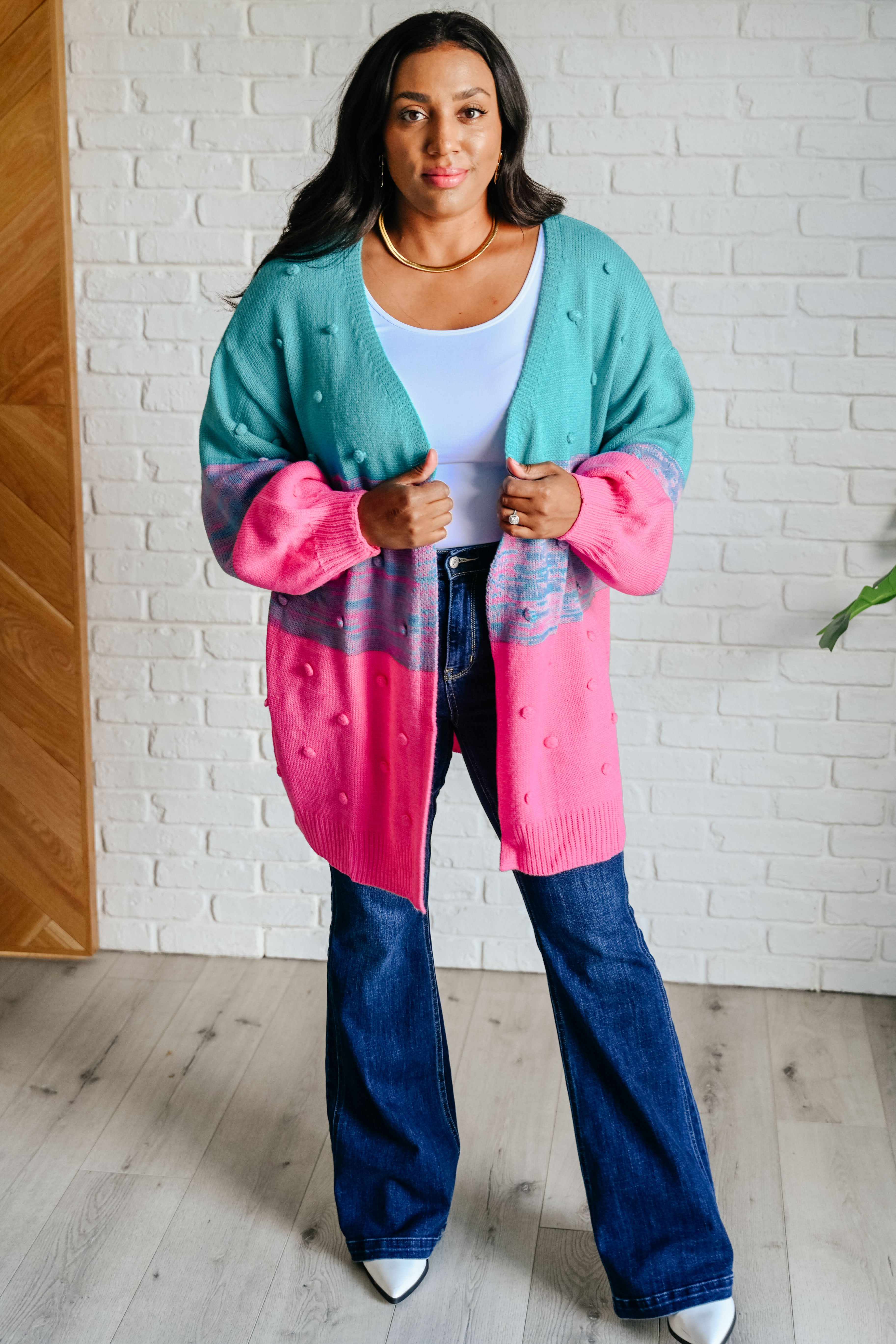 Woman wearing teal and pink oversized cardigan with textured dots layered over white top and flared jeans indoors