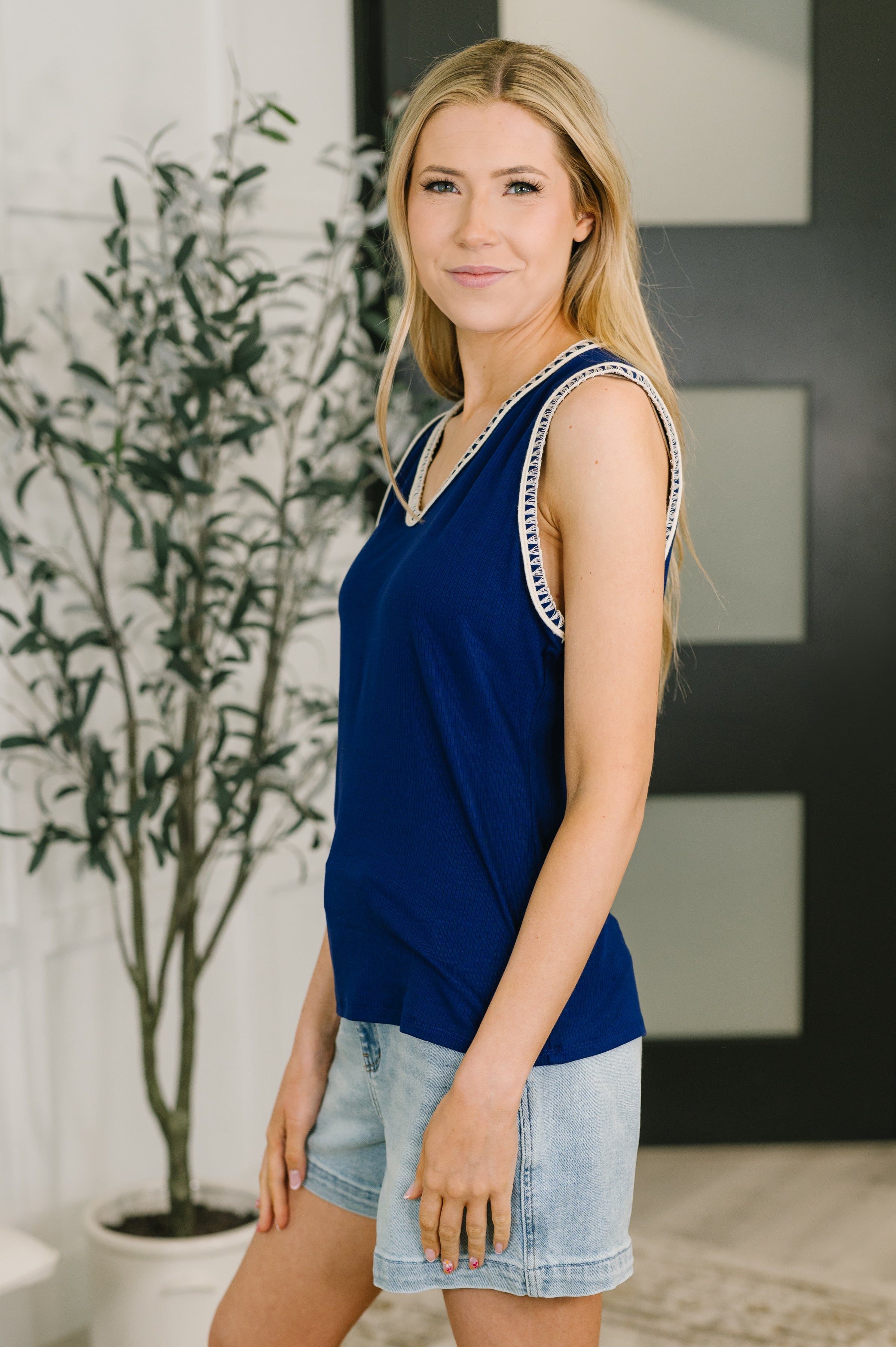 Woman wearing navy sleeveless V-neck tank with contrast trim paired with light denim shorts in bright indoor setting