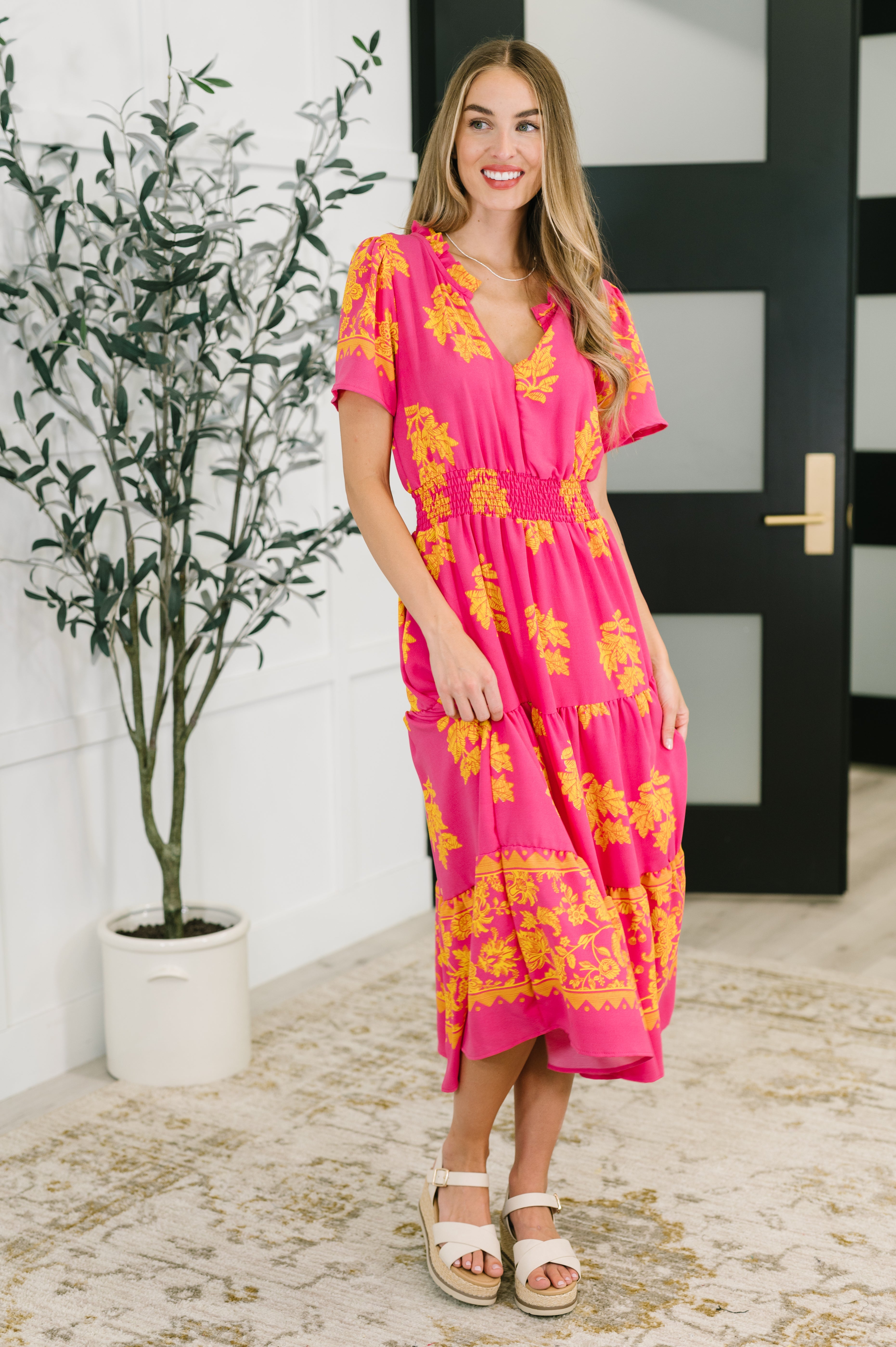Woman indoors wearing fuchsia V-neck dress with yellow floral design and smocked waist standing near plant
