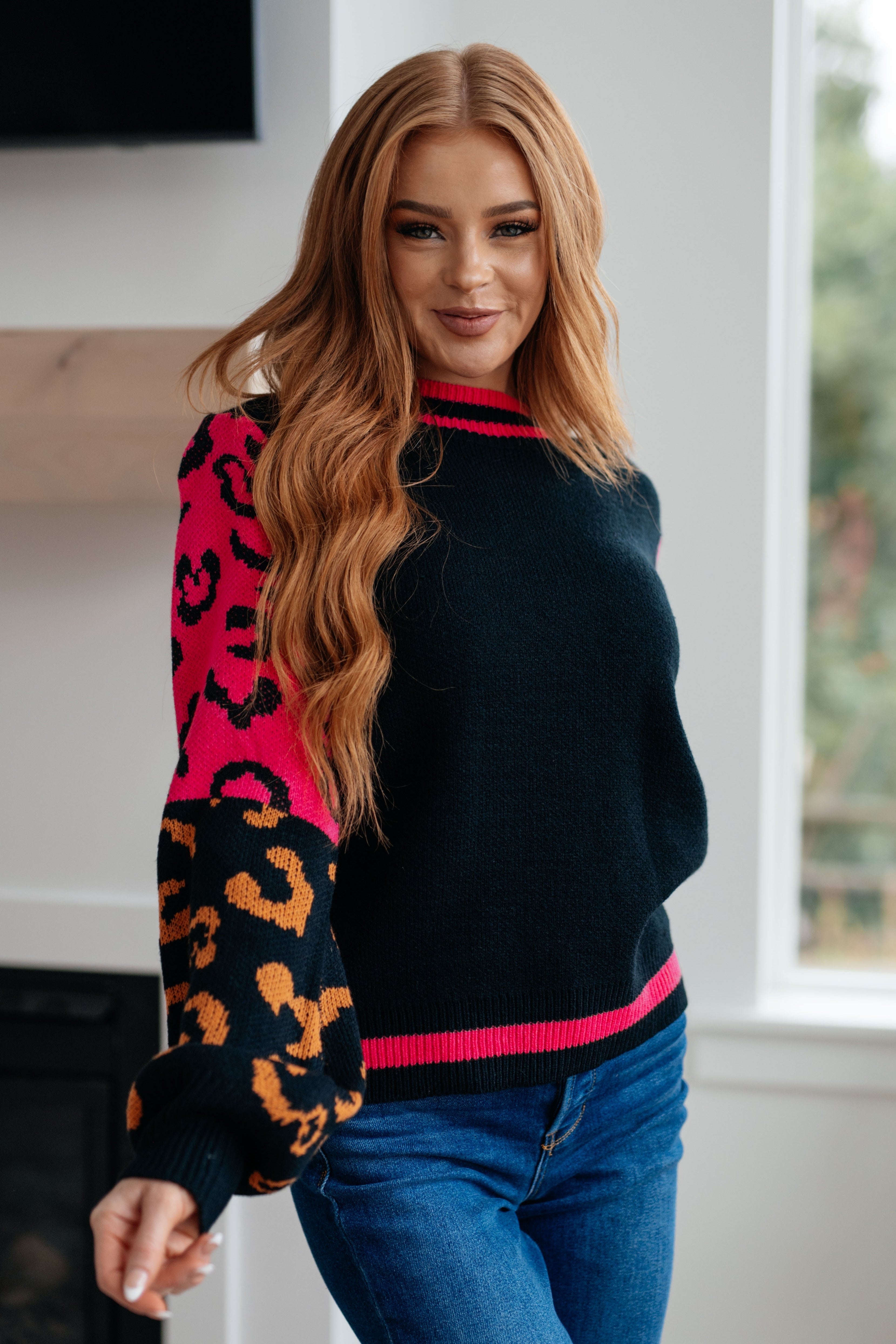 Woman wearing black sweater with contrasting pink and orange animal print sleeves posing indoors near window