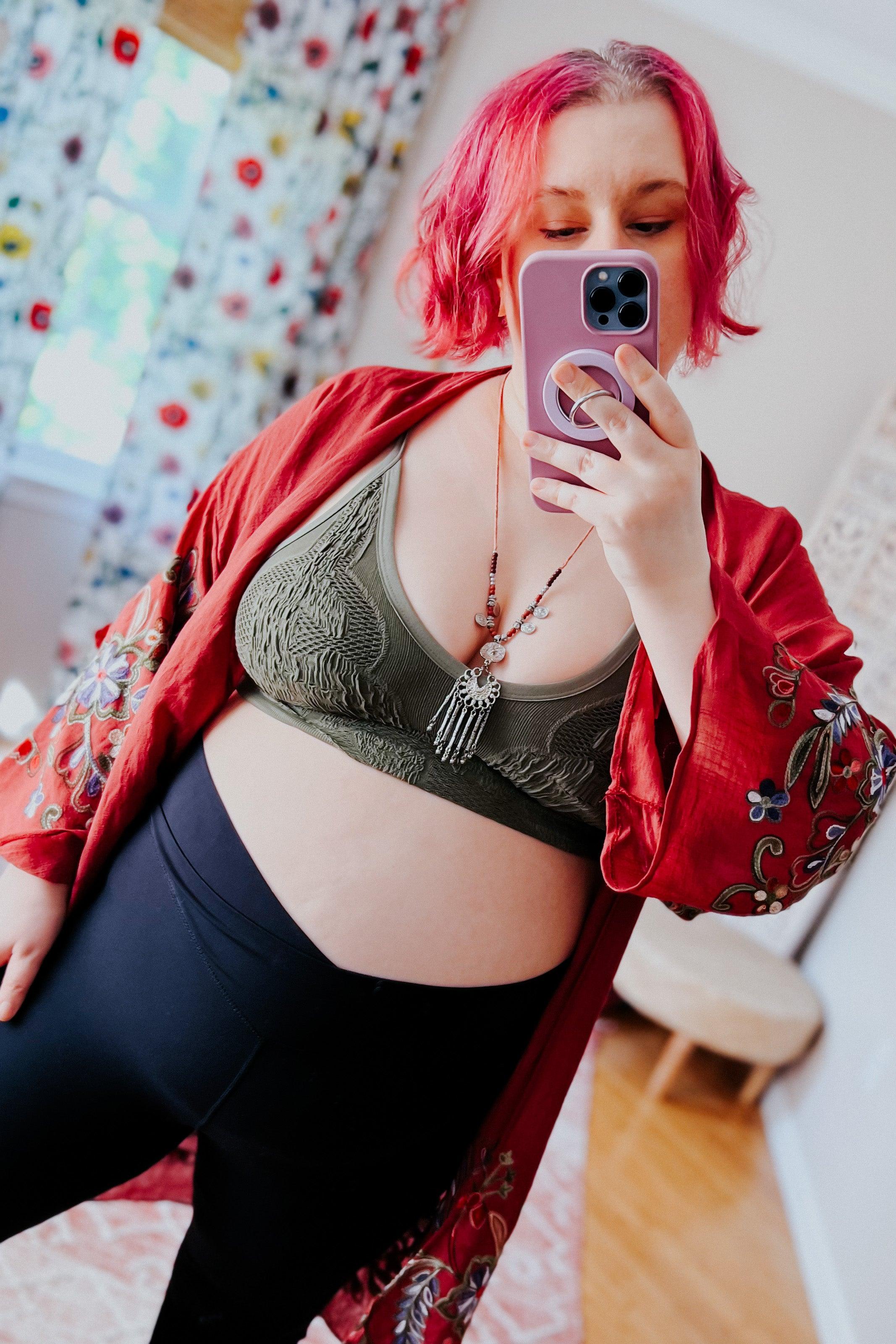 Woman with bright pink hair wearing an olive textured brami and black leggings taking a selfie indoors