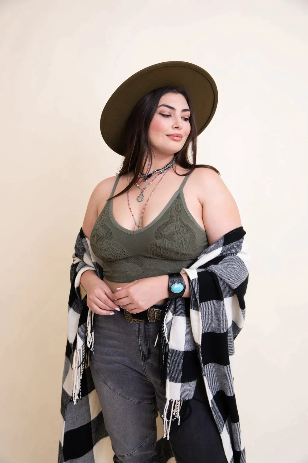 Woman wearing olive textured brami and layered necklaces with wide-brimmed hat and checkered shawl indoors