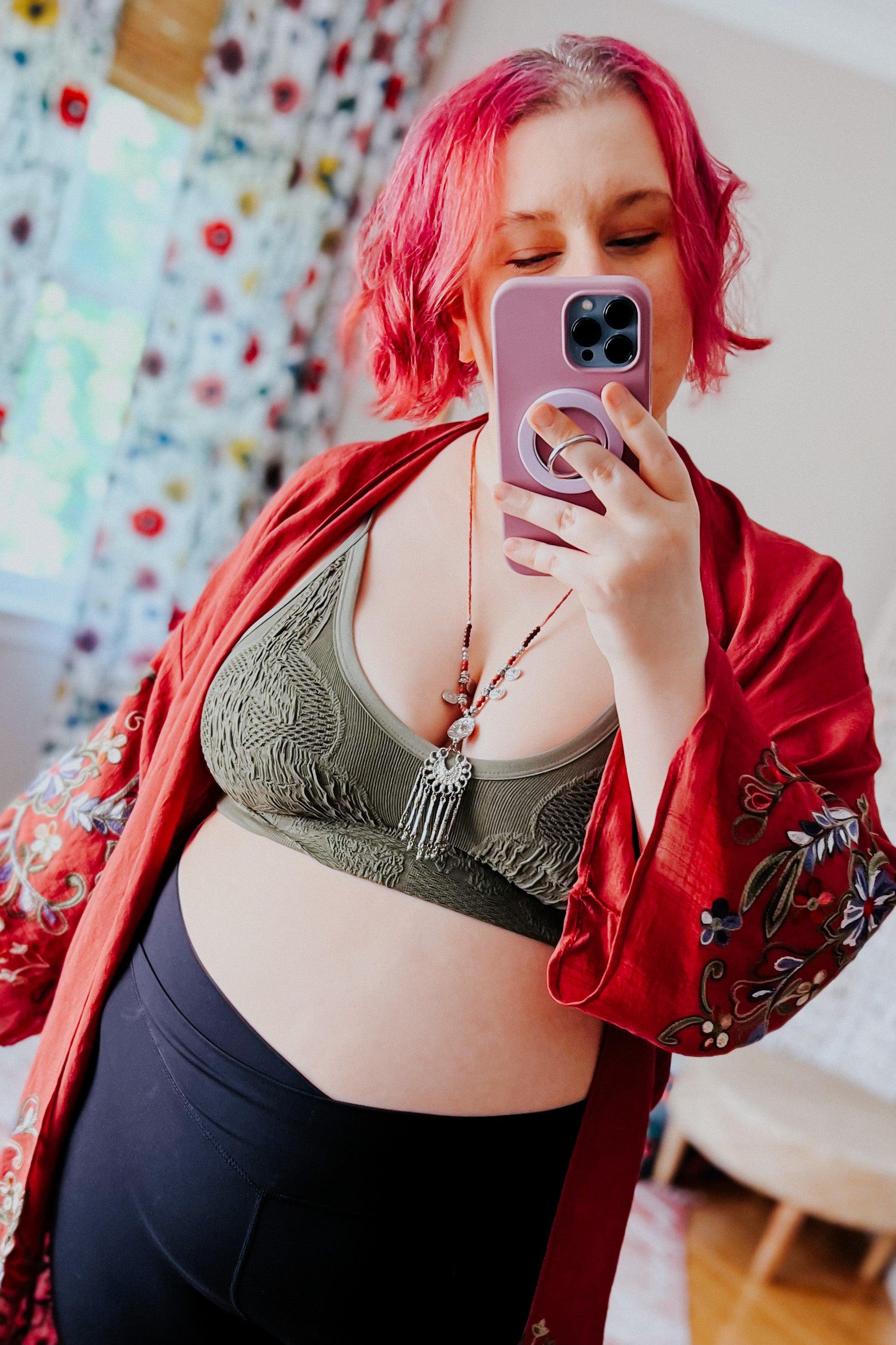 Woman with bright pink hair wearing olive textured brami and patterned red robe taking mirror selfie indoors
