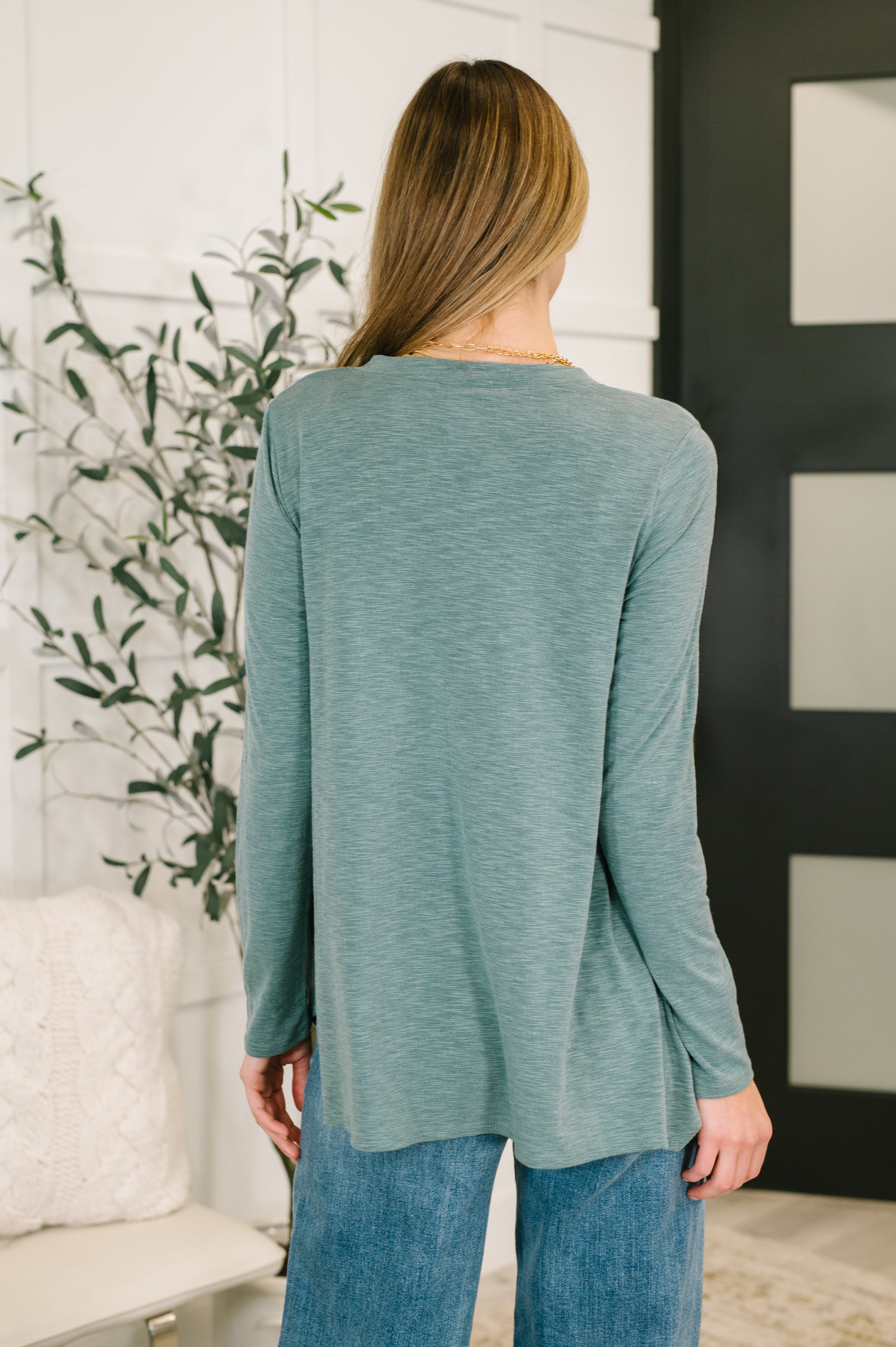Woman wearing a light teal v neck long sleeve top and blue jeans standing in an indoor setting.