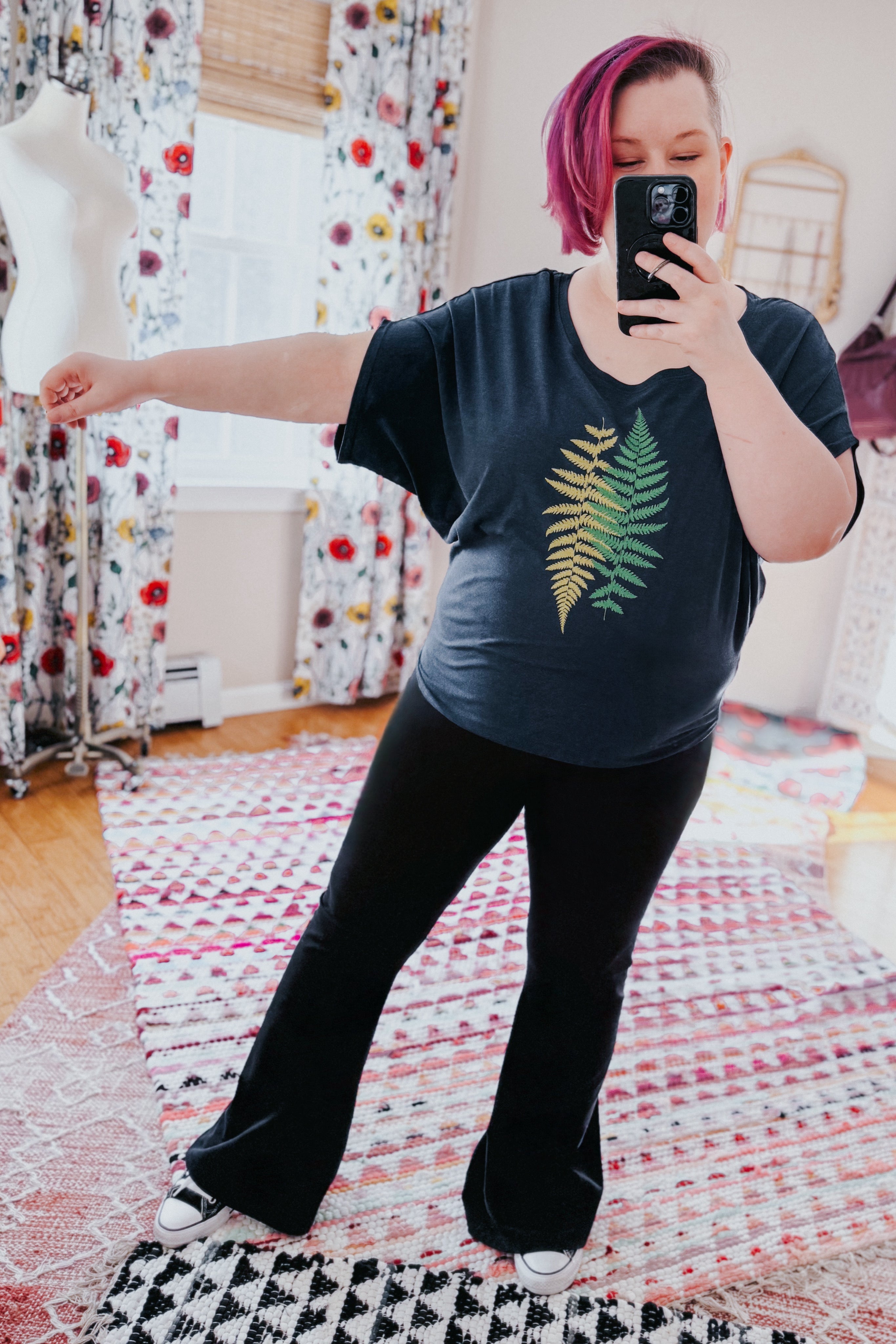 Woman with short pink hair wearing navy bamboo dolman top with fern design and black pants taking mirror selfie indoors