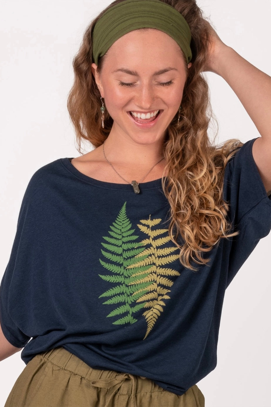 Smiling woman wearing navy bamboo dolman top with green and yellow fern print, olive headband, and olive pants