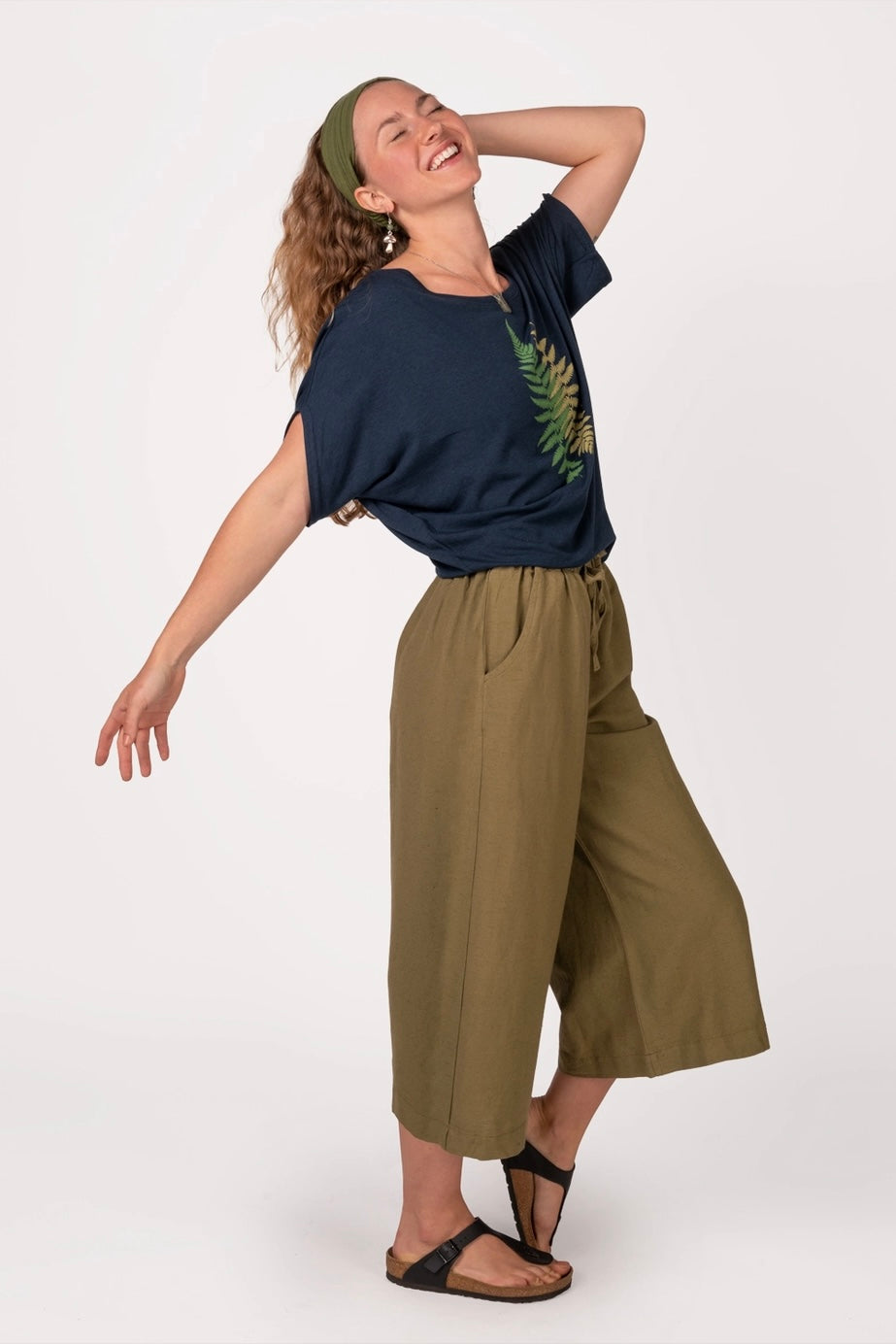 Woman joyfully posing wearing navy bamboo dolman top with fern print, olive wide-leg pants, and black sandals
