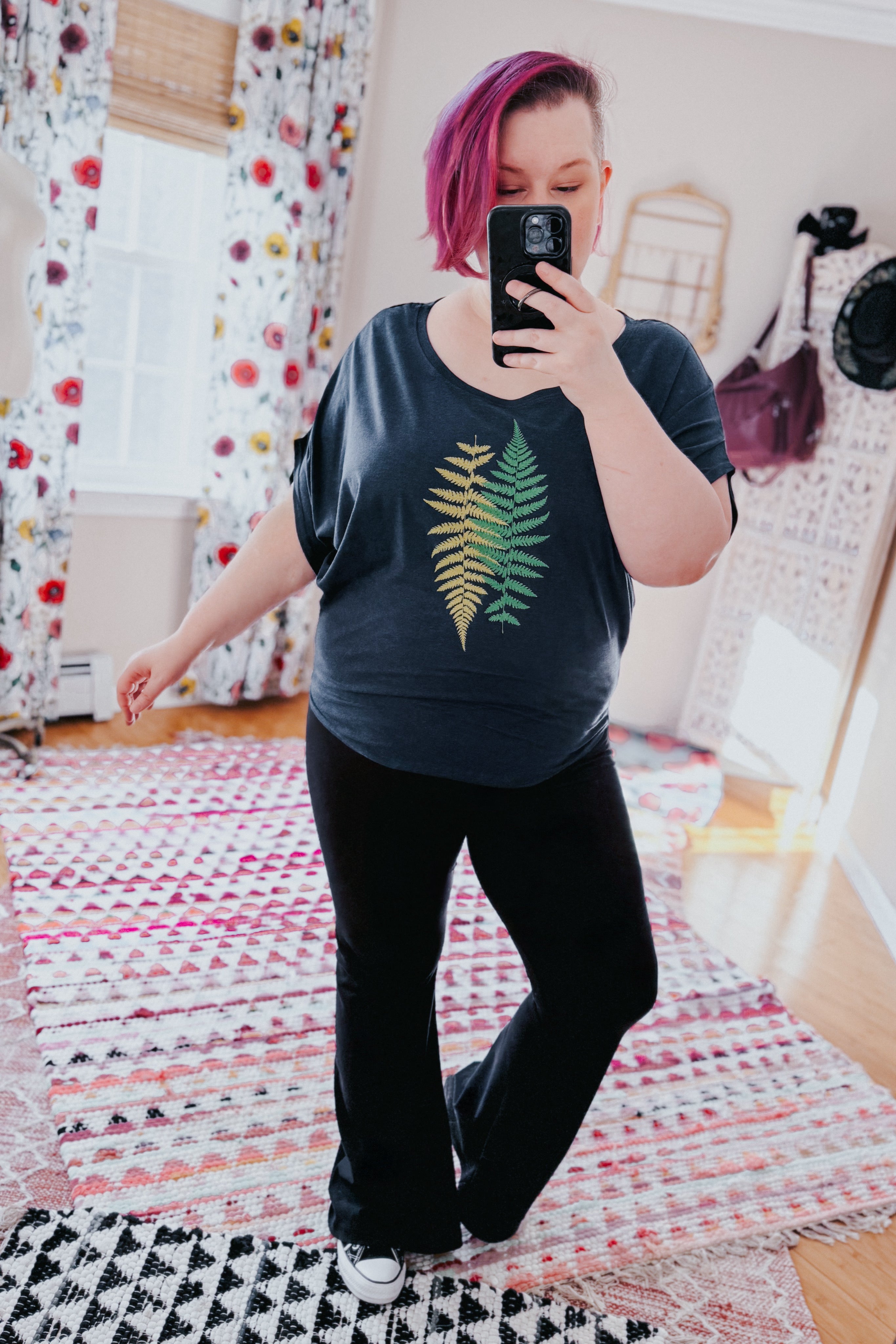 Woman with pink hair wearing navy bamboo dolman top with fern print and black pants taking mirror selfie indoors