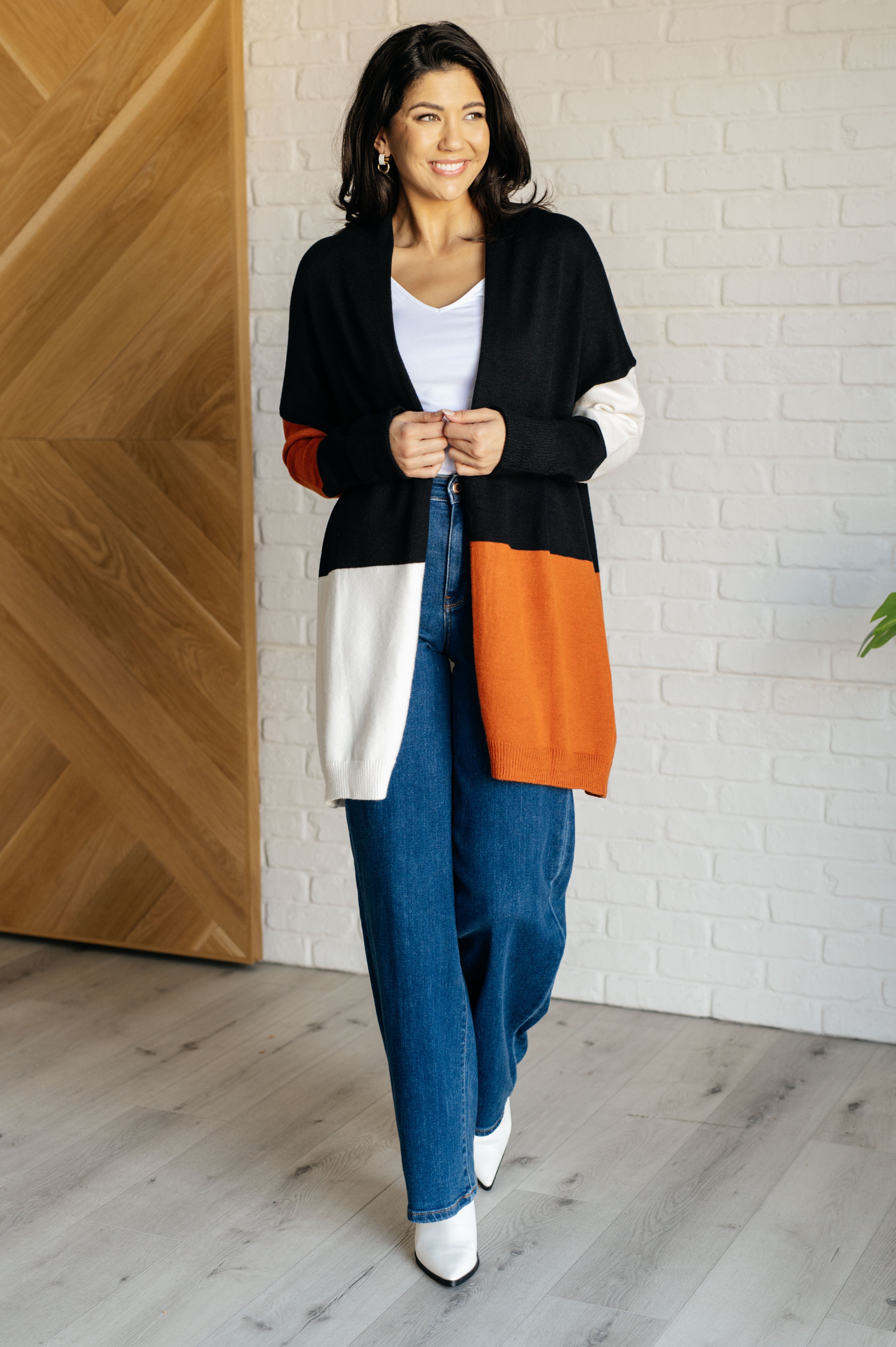 Smiling woman wearing black, white, and orange color block open front cardigan with blue jeans and white boots indoors