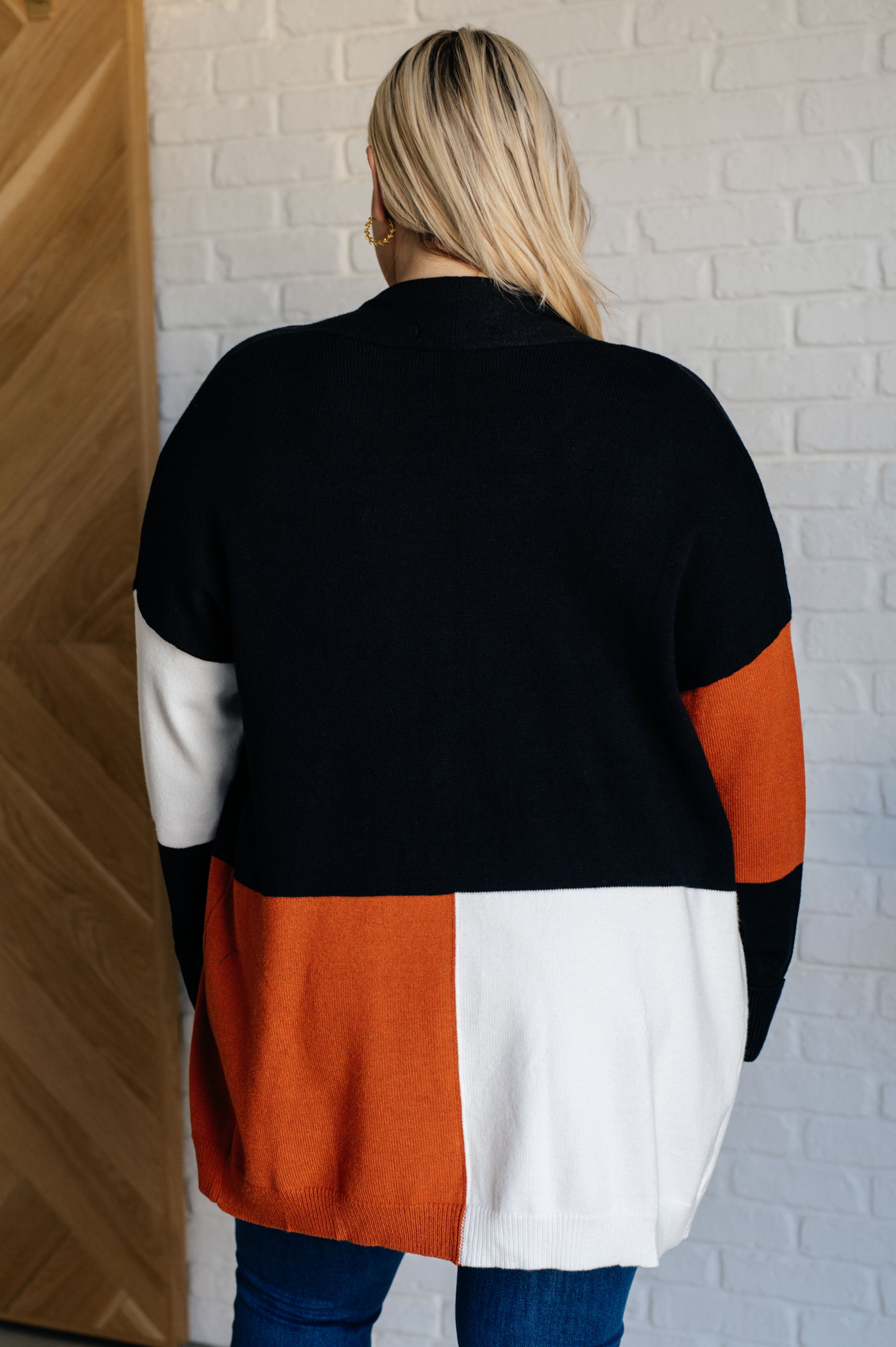 Woman wearing color block open front cardigan in black, white, and burnt orange standing indoors facing away