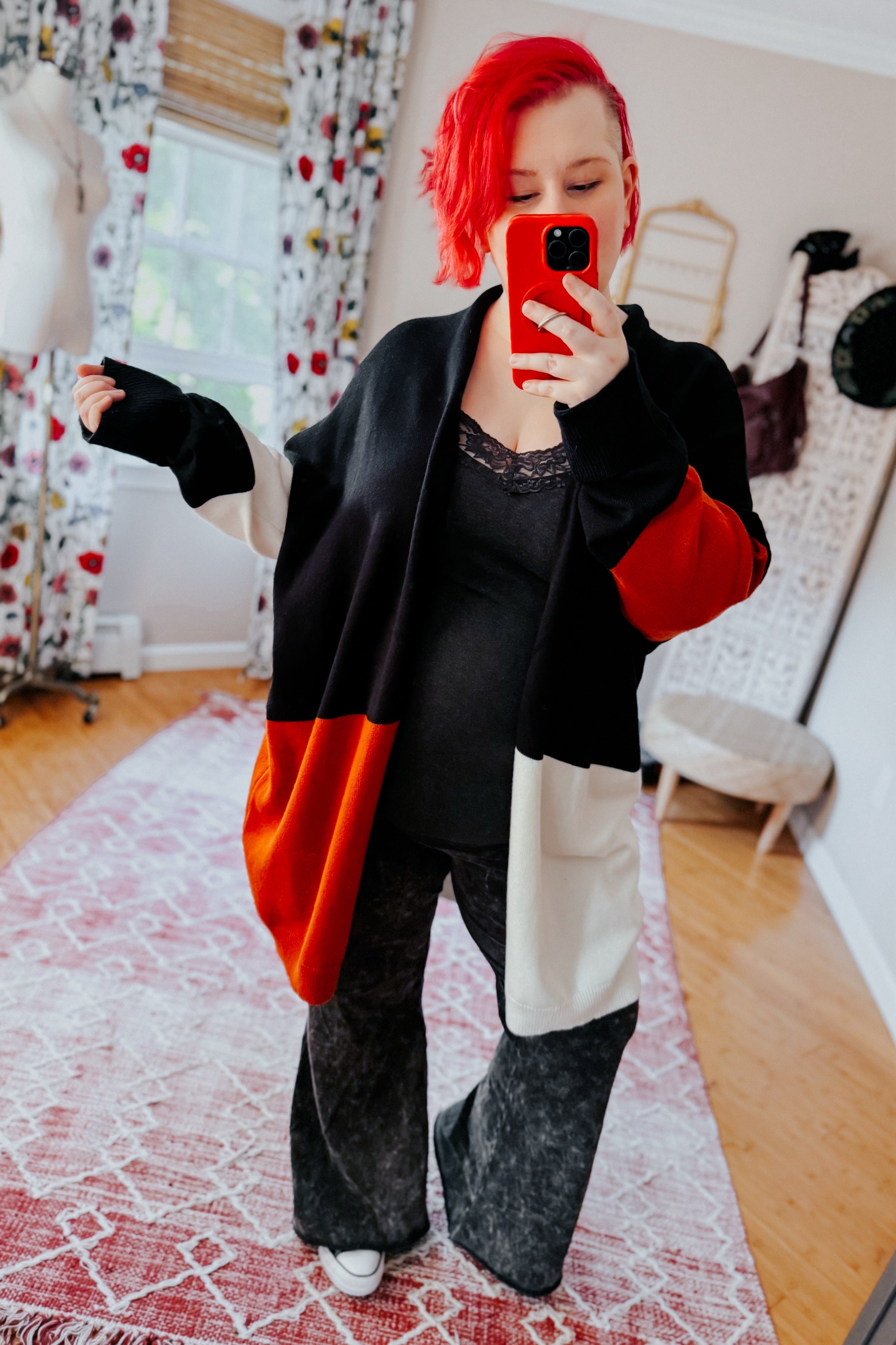 Woman with bright red hair wearing color block open front cardigan in black, red, and white taking mirror selfie indoors