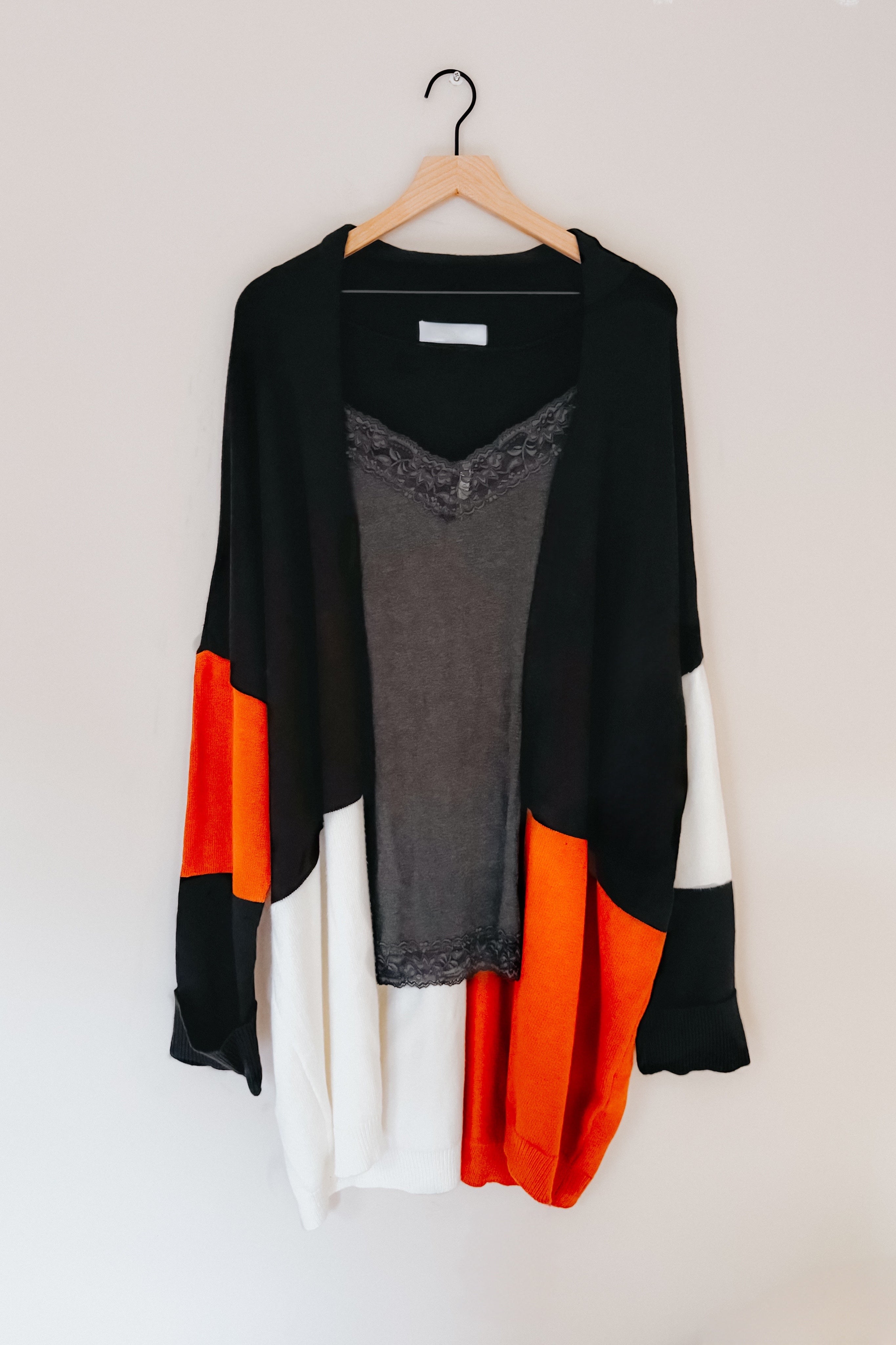 Black open front cardigan with orange and white color block sleeves layered over gray lace-trim tank top on wooden hanger