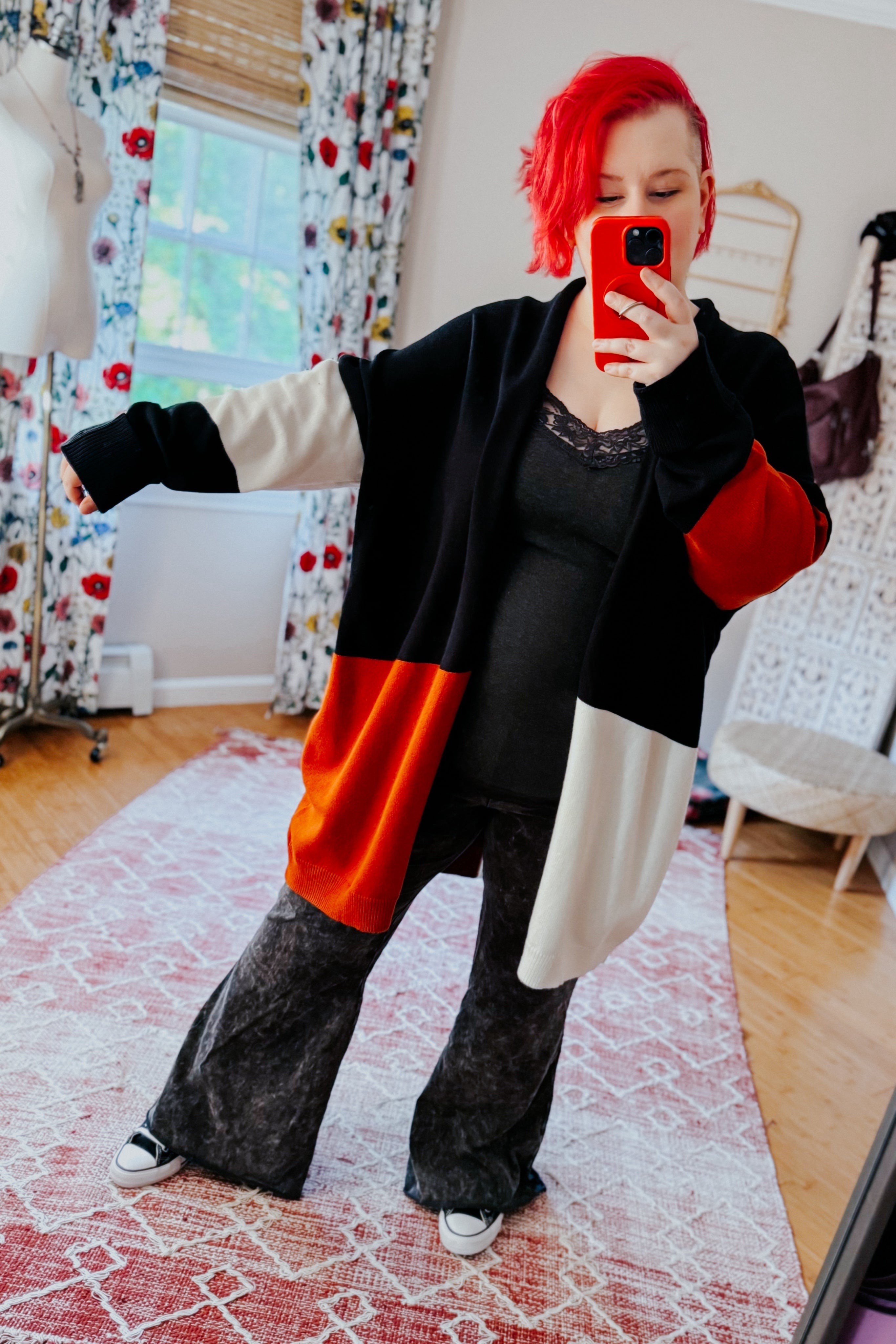 Woman with red hair wearing color block open front cardigan in black, cream, and rust posing in bright room with patterned curtains